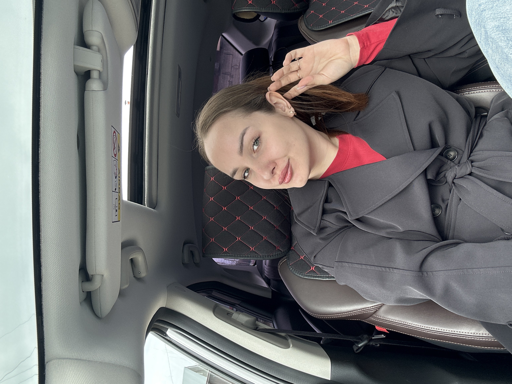 A woman is sitting in the passenger seat of a car. She is wearing a red top and a gray trench coat. She has brown hair and is smiling at the camera. The car's interior is light gray, with black seat covers embroidered with red diamonds. The image was taken in Chaikovsky, Russia.