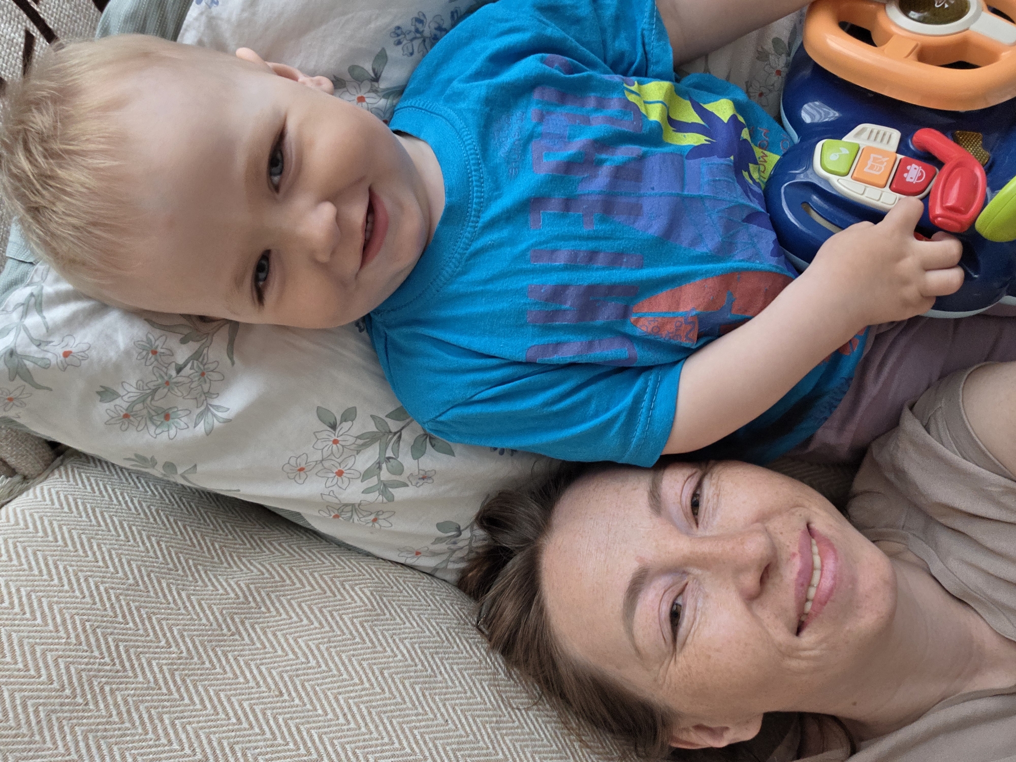A top-down shot features a young child and an adult, possibly a parent, lying down together on what appears to be a bed or sofa. The child, with blond hair and a blue shirt, is holding a blue and orange toy steering wheel with colorful buttons. The adult, likely a woman, is smiling at the camera, lying parallel to the child. The scene is indoors, and the background shows a pillow with a floral pattern and a textured beige surface. There is no indication of location to determine the city of Chaikovsky, Russia.
