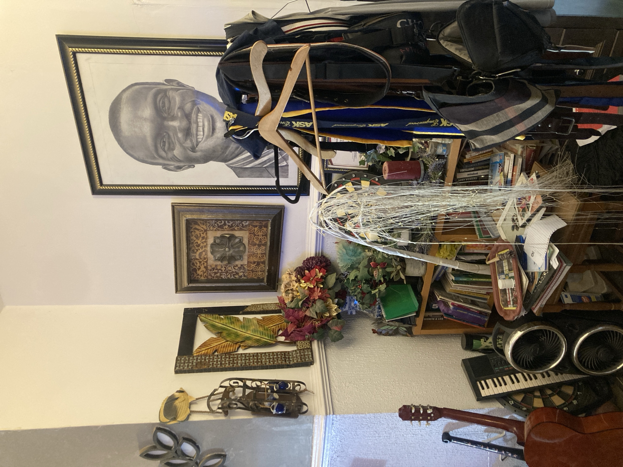 The image depicts an interior scene. Dominating the left wall is a framed pencil portrait of a smiling man. Below it are several framed artworks: a square floral design, a leaf-themed picture, and a metal decorative piece. A wooden bookshelf stuffed with books and various items stands against the right wall. An item of clothing hangs from the shelf, and there are floral decorations on top of it. A dartboard, keyboard, and guitar can be seen at the bottom. The location is uncertain, as there is no clear indication of Jos, Nigeria.