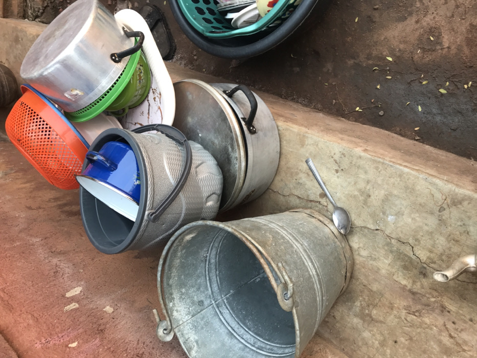 A variety of containers, including pots, baskets, and a metal bucket, are piled on a concrete surface. The pots, made of silver-colored metal, are stacked with lids askew. Bright orange and green baskets are visible. A blue pot sits nested within a larger gray bucket with a black handle. A metal spoon rests precariously on the rim of the metal bucket, which leans against a concrete wall. A tap fixture is on the right side of the image.