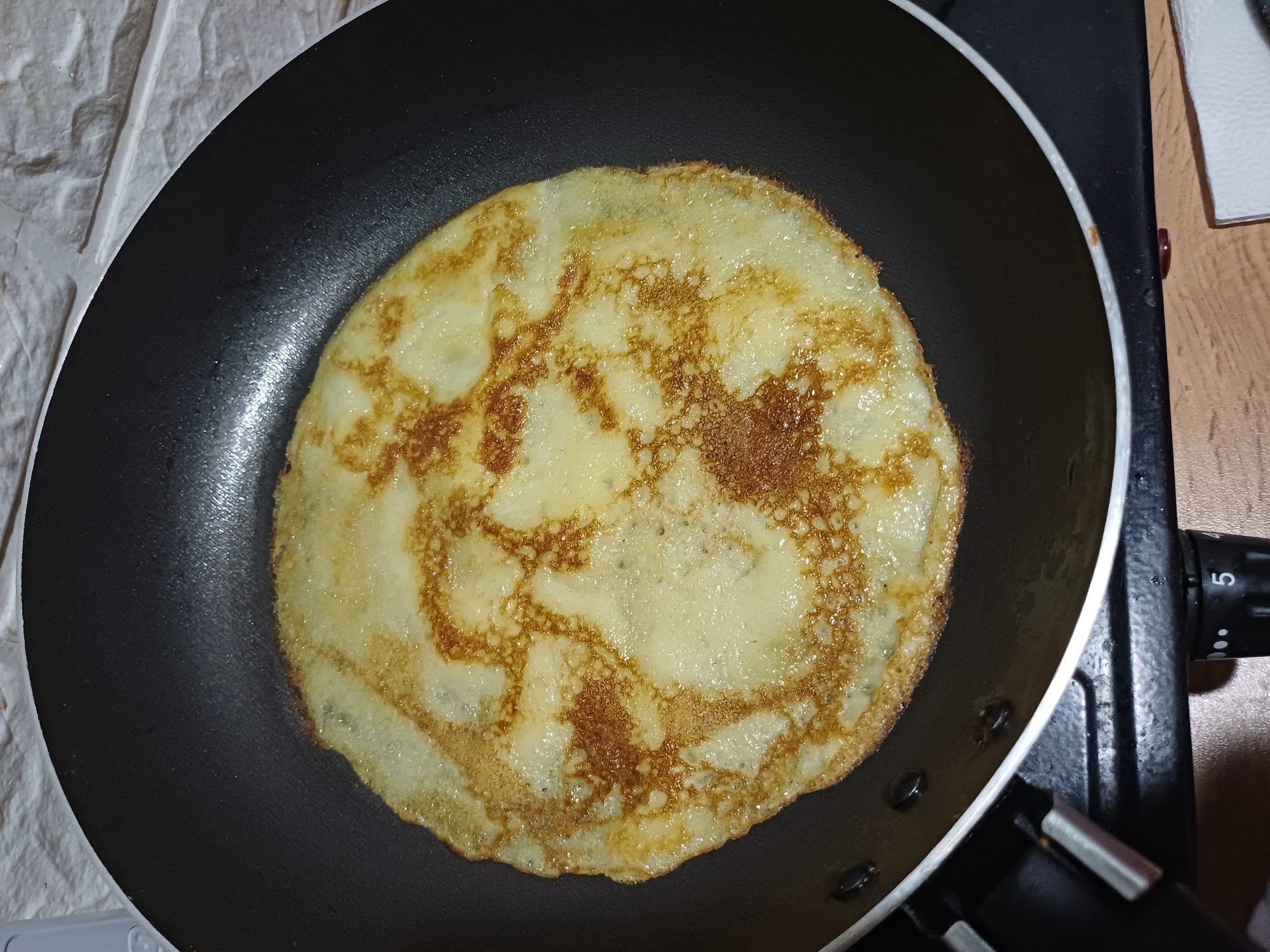A close-up, overhead shot captures a single, golden-brown pancake being cooked in a black non-stick frying pan. The pancake, circular and thin, exhibits a slightly uneven texture with darker brown, crispy edges and lighter, more translucent sections in the center. Small air bubbles are visible on its surface. The frying pan is placed on what appears to be a stovetop, indicated by a black knob with a white "5" marking on its right side, suggesting a temperature setting. The background on the left shows a textured, white wall, possibly made of brick or stone. To the right of the pan, a wooden surface is visible, along with a white rectangular object. The lighting suggests an indoor setting, possibly a kitchen. The image focuses solely on the cooking pancake, with no people or other food items visible. The overall impression is that of a home-cooked meal in progress.