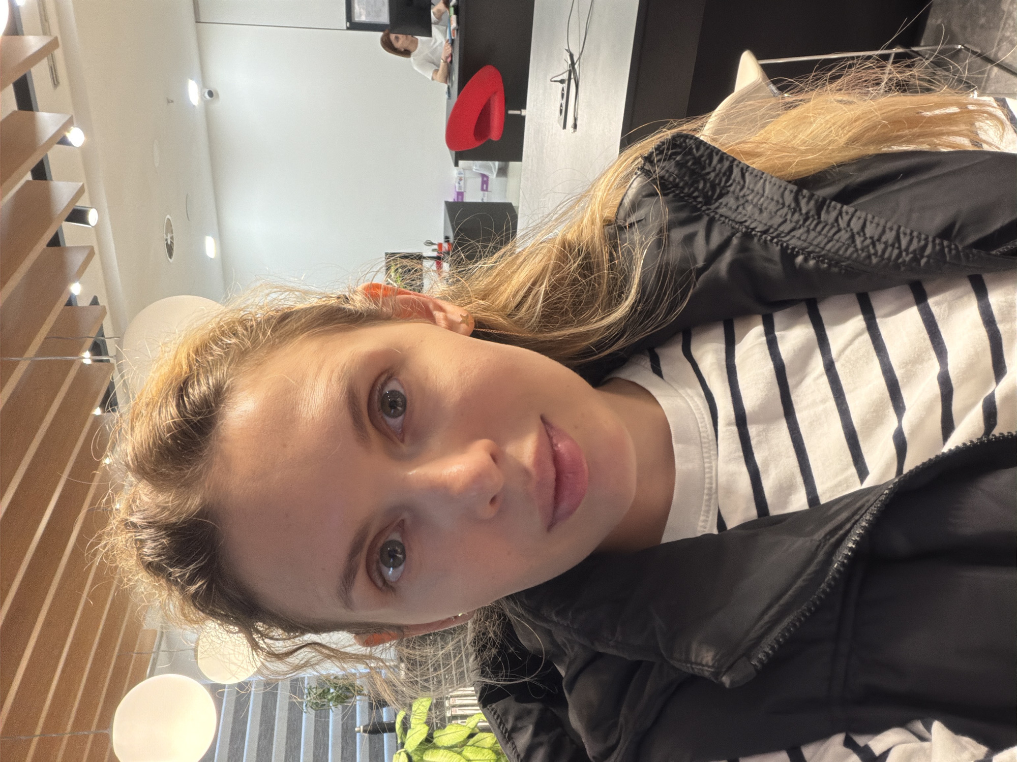 A woman is taking a selfie indoors. She is wearing a black puffer jacket over a white and navy striped shirt. The location appears to be a bright, modern interior, possibly an office space. In the background, there is a glimpse of someone seated at a desk and a red office chair. The image may have been taken in Podgorica, Montenegro.