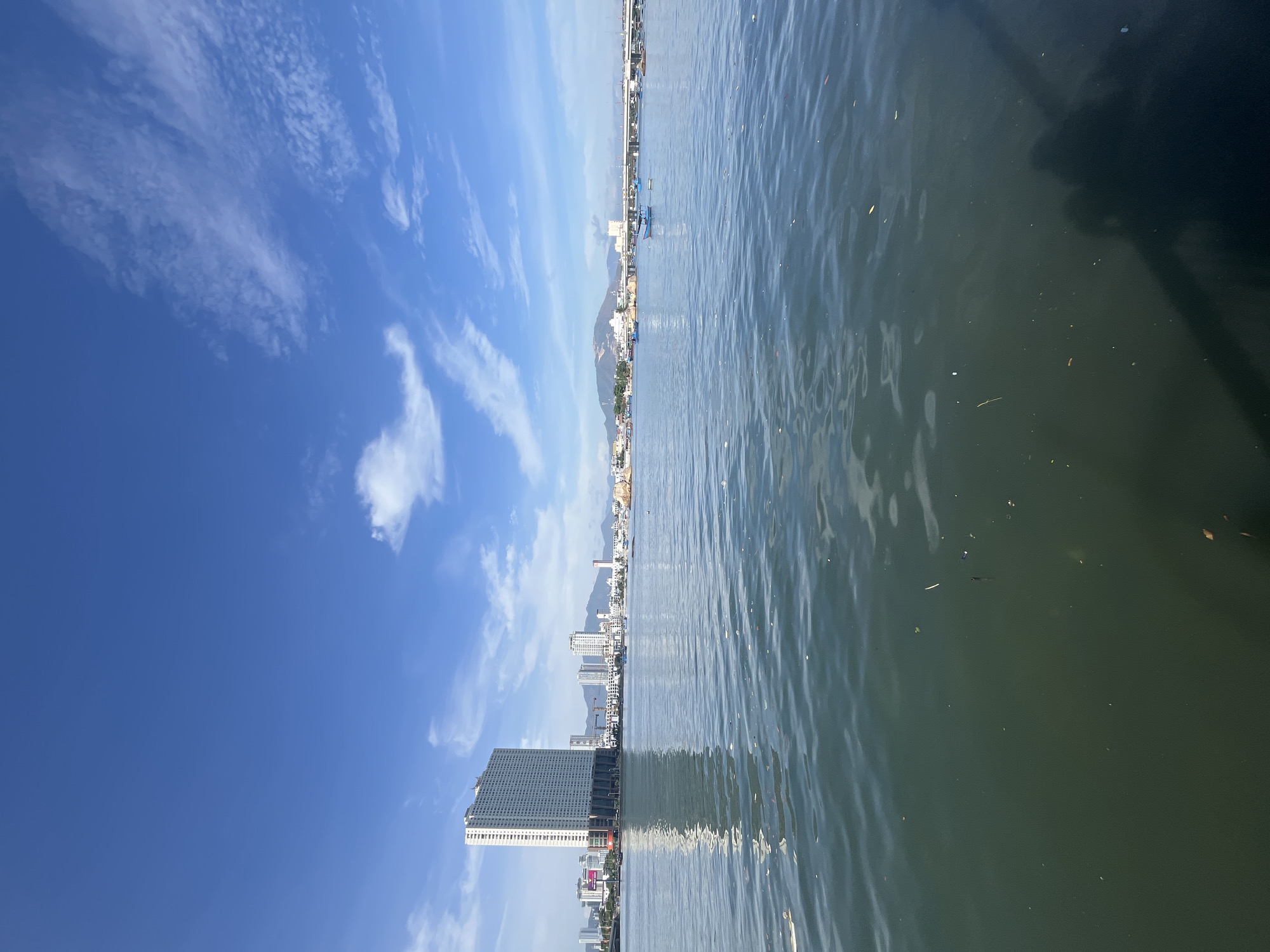 The image depicts a cityscape reflected in a body of water, likely taken from a boat in Bac Nha Trang Ward, Vietnam. A tall, modern building with a patterned facade stands prominently at the bottom left. Other buildings, including high-rises and smaller structures, line the horizon. A mountain range is visible in the distance. The sky is bright blue with scattered white clouds. The water is rippled and reflects the sky and buildings. Some debris is floating in the water. A dark shadow appears in the upper right corner, possibly from part of the boat or camera.