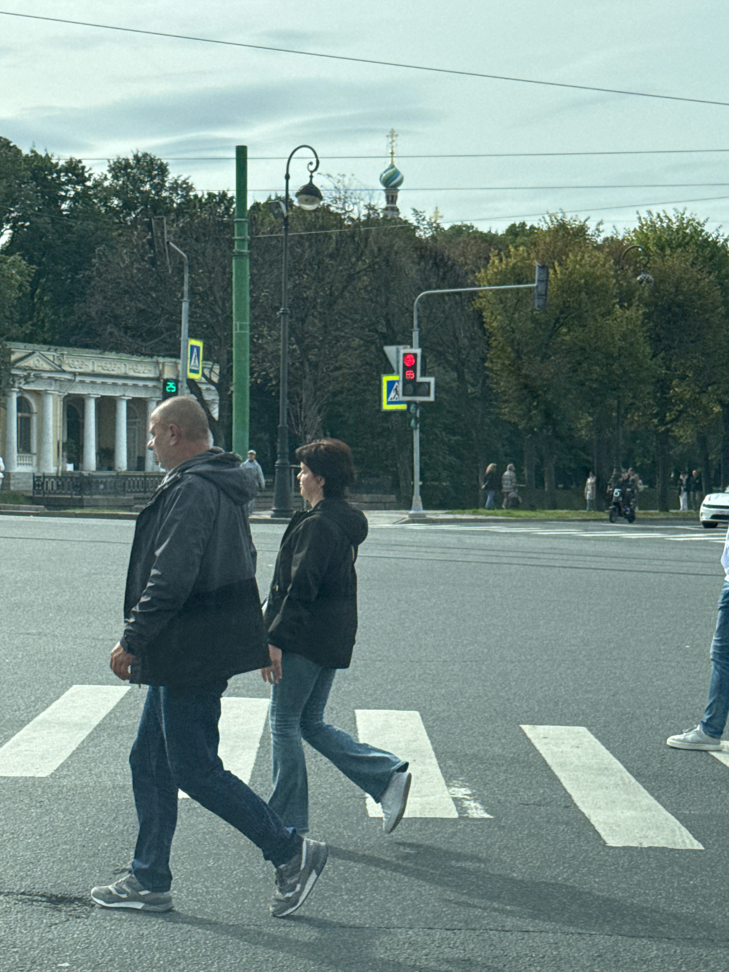 The image depicts a street scene in Saint Petersburg, Russia, featuring a crosswalk with three individuals walking across it. The sky is overcast. In the background, trees and a building with classical architecture are visible. A traffic light displaying a red signal is also present. The foreground shows the asphalt road and white crosswalk lines.
The central figures are two adults, a man and a woman, walking side-by-side. The man, on the left, is wearing a dark jacket, blue jeans, and grey sneakers. He is bald. The woman, to his right, is wearing a black jacket and blue jeans. Behind them, partially visible on the right edge of the frame, is a third person wearing blue jeans and white sneakers.
The architectural style of the building in the background, with its colonnade and pediment, suggests a historical or neoclassical design common in Saint Petersburg. The presence of trees indicates a park or a tree-lined avenue, also characteristic of the city. The traffic light confirms an urban setting with regulated pedestrian and vehicular traffic. The overall atmosphere is one of everyday city life under a subdued sky.