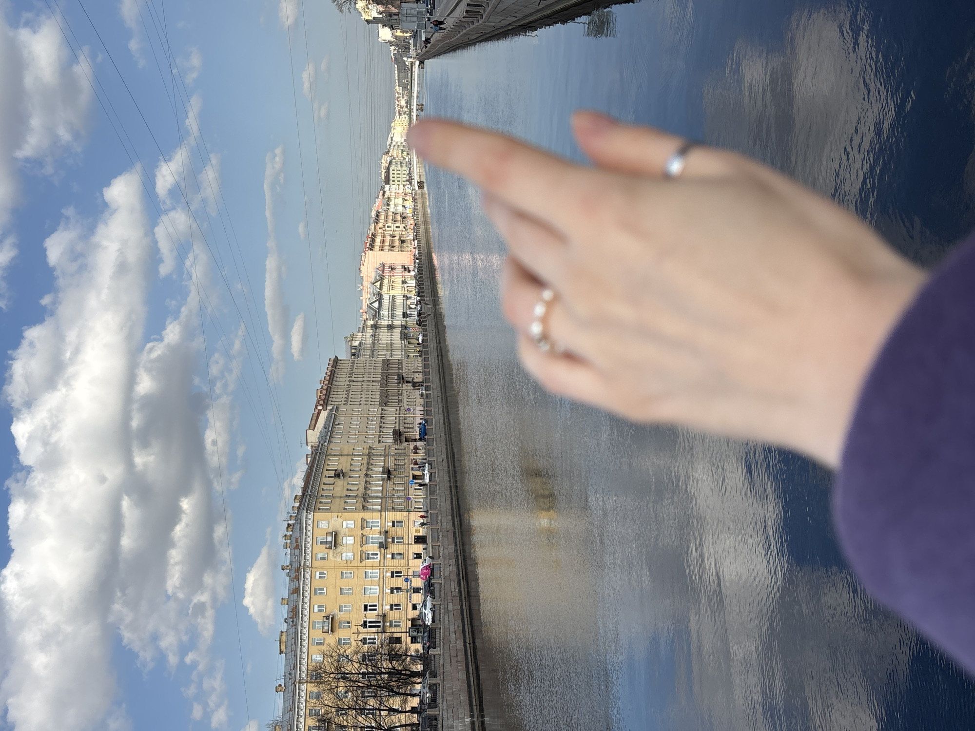 The image presents an urban landscape along the Fontanka River in Saint Petersburg, Russia, under a clear sky with scattered cumulus clouds.
In the immediate foreground, a blurred, light-skinned human hand is prominent, partially obscured by a dark purple garment at the bottom left. The hand wears a silver-colored band on the ring finger and a ring adorned with small white spherical beads on the index finger. The index finger is extended, pointing diagonally towards the upper right, into the mid-distance along the river.
The Fontanka River occupies the central and lower portions of the scene, its surface reflecting the blue sky and white clouds with visible ripples. Granite embankments line both sides of the river.
On the left bank, beyond the embankment, a street is visible with parked vehicles, including a bright pink car. Bare deciduous trees are interspersed along the street. Behind these, a continuous row of multi-story classical buildings extends into the background. The closest prominent building is pale yellow with numerous windows and small balconies.
On the right bank, a promenade or street runs along the embankment, followed by a consistent line of multi-story buildings, presenting facades in pastel shades of pink, cream, and white, also stretching into the distance. Pedestrians are discernible on both banks in the background, and limited vehicular traffic is present on the streets.
Multiple arched bridges span the river in the far distance, gradually receding from view. The upper third of the image is dominated by a bright blue sky dotted with large white clouds. Several thin, dark overhead lines, likely power lines, crisscross the sky horizontally above the distant cityscape. The entire scene is illuminated by direct sunlight.