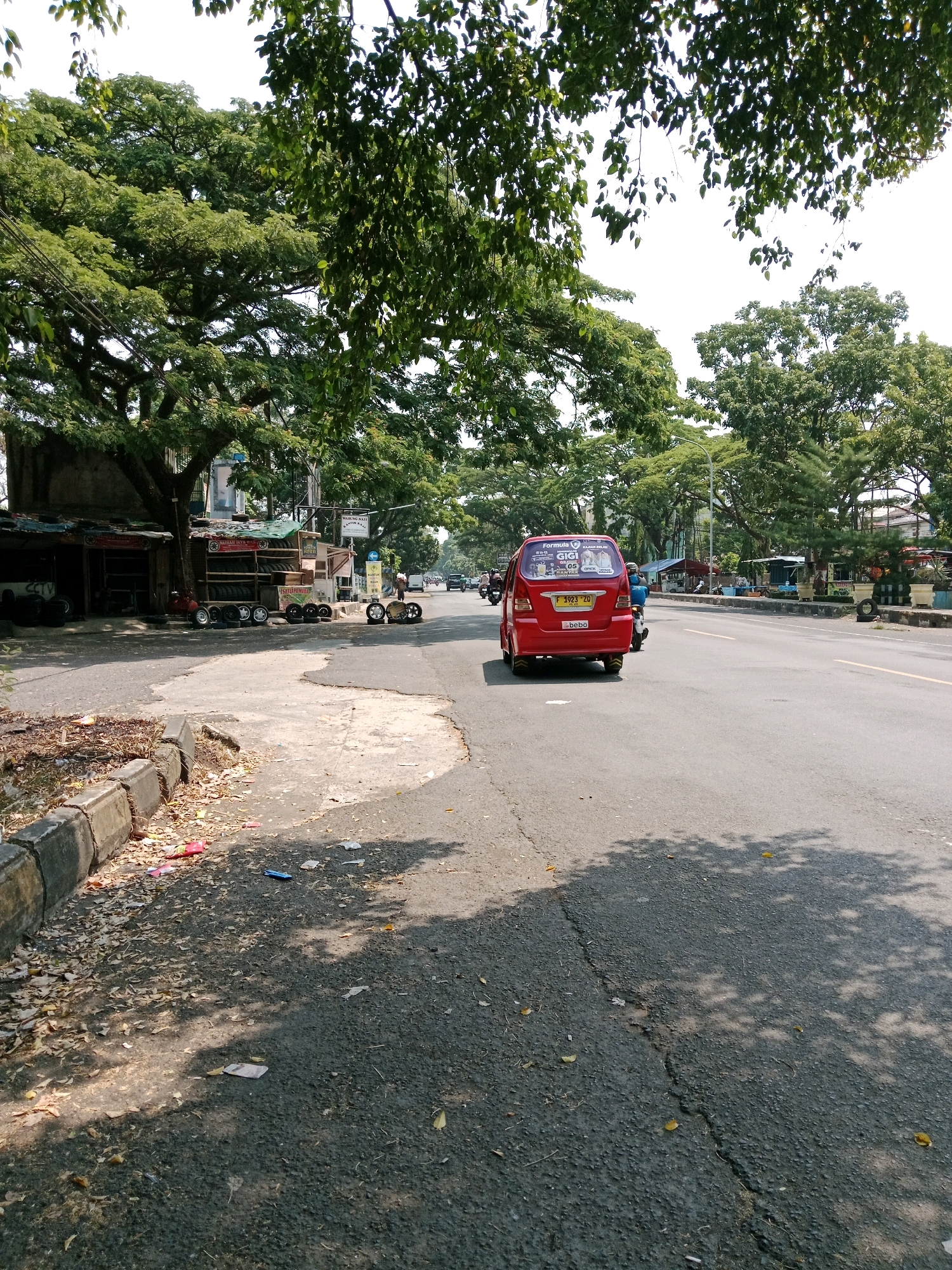 The image shows a street scene in Karangtengah, Indonesia, on a sunny day. A red minivan, identified by the license plate "F 3923 ZD", is traveling away from the camera on a paved road. The minivan has stickers on its rear window. To the left of the road, there is a sidewalk with a concrete curb. Along the sidewalk, there are several small shops selling tires. One shop has tires displayed on racks, and there are also loose tires and wheels on the ground. A person is visible near one of the shops. Further down the road, more shops and trees line the street, with other vehicles and people in the distance. The ground near the curb is littered with leaves and some trash. The scene is dominated by the shadows of trees cast on the road and sidewalk, indicating the time of day.