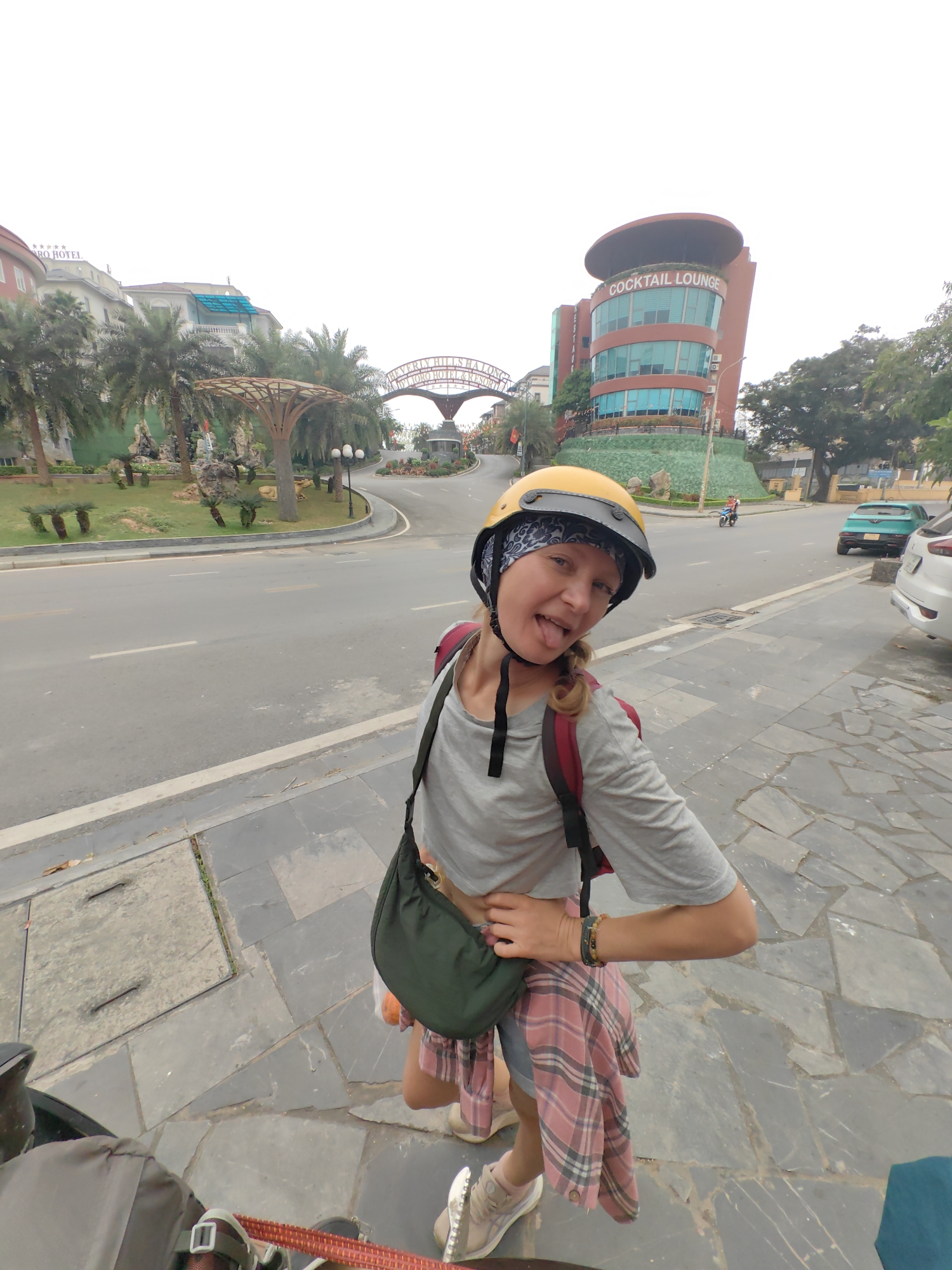 A woman in a yellow helmet and patterned head covering is standing on a gray sidewalk next to a gray road, sticking out her tongue at the camera. She is wearing a gray t-shirt, a red and white plaid shirt tied around her waist, and has a green purse across her chest. Buildings, including one marked "COCKTAIL LOUNGE," are visible across the road. The location is Bai Chay Ward, Vietnam. A motorcycle and two cars are visible on the road. There are palm trees in the background.