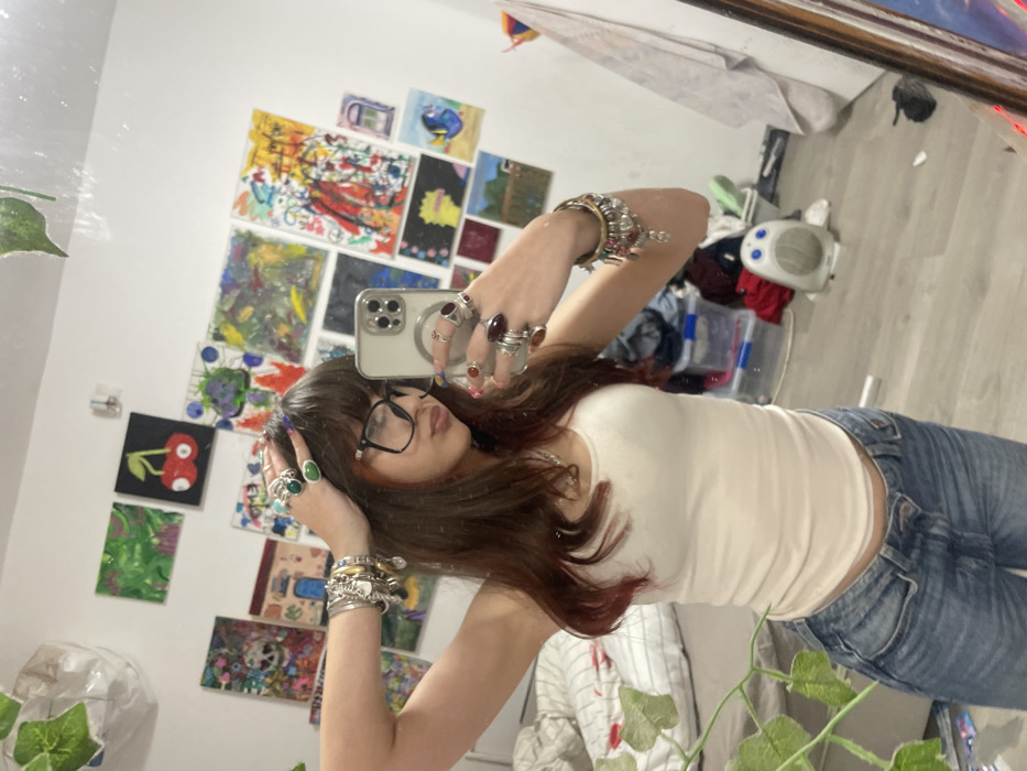 A young woman takes a selfie in a mirror. She wears eyeglasses, a white tank top, and blue jeans. Her brown hair has red highlights. She accessorizes with many rings and bracelets. Behind her are various paintings, presumably her own artwork, hanging on the wall. The room appears cluttered, with items on the floor and a plant in the foreground.