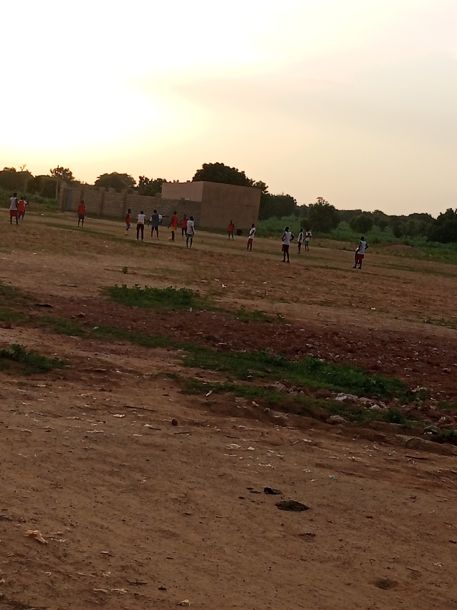 A group of individuals, likely boys, are playing a game of soccer on a dirt field at dusk in Yardaje, Nigeria. The sky is a hazy orange and yellow, indicative of sunset. In the background, a low brick wall surrounds a building, suggesting a residential area. Trees are visible behind the wall, and more trees and vegetation line the horizon. The field itself is uneven and appears to be a mix of dirt and sparse grass. The individuals are dressed in casual clothing, with some wearing t-shirts and shorts. They are scattered across the field, some in motion, while others appear to be standing or observing. The overall atmosphere is one of informal recreation in a rural or semi-rural setting.