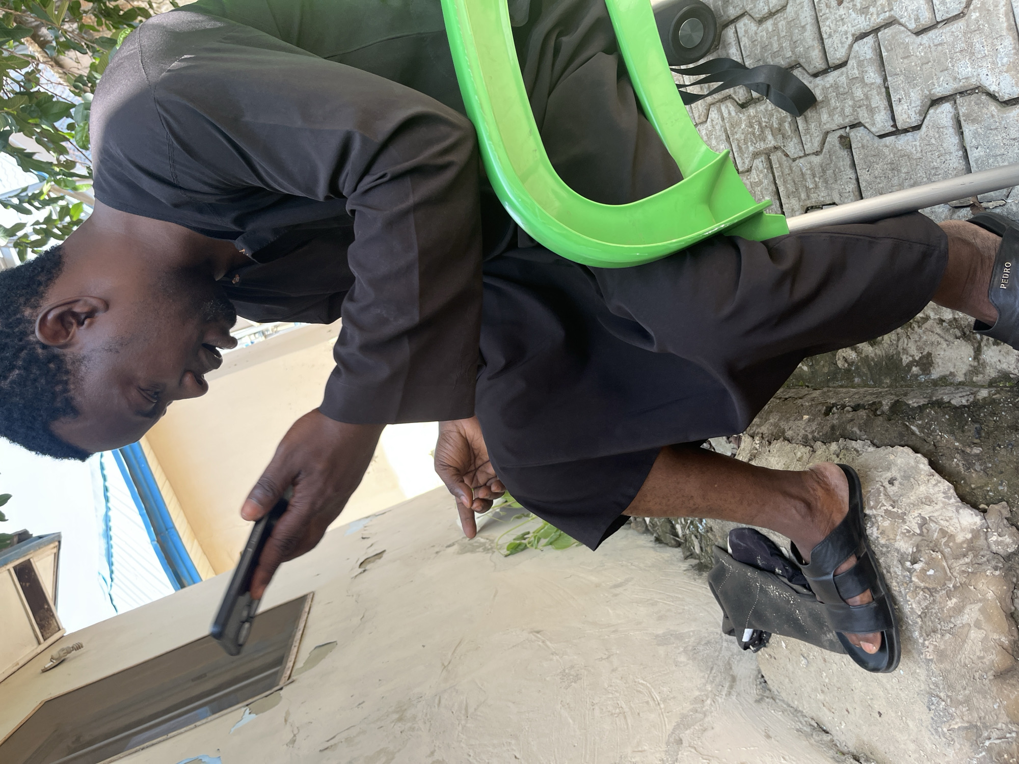 A man wearing a black robe and black sandals is sitting in a green plastic chair. He is holding a black smartphone in his right hand and appears to be looking at it. His left hand is resting on his leg. The scene is outdoors, with a textured grey pavement beneath the chair and a light-colored wall in the background. There is some green foliage visible. The image is taken from a low angle, looking up at the man. The sunlight suggests it is daytime. The setting appears to be a residential or outdoor area in Galadimawa Village, Nigeria. The man's expression is neutral. Visible text on the sandals reads "PEDRO".