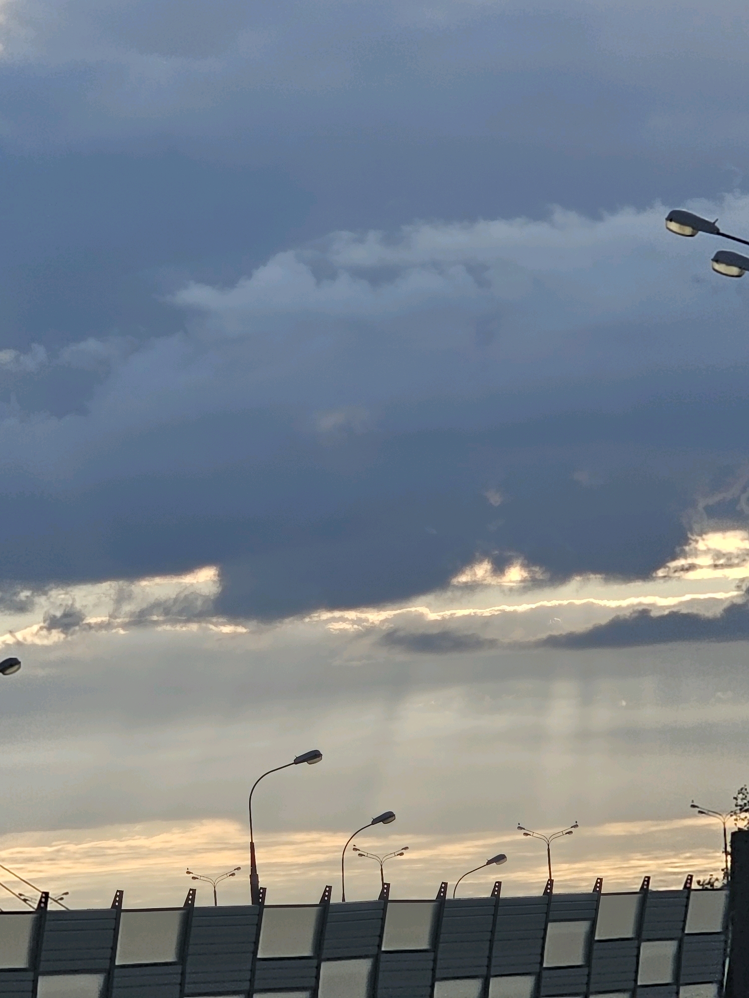 The image depicts a cloudy sky with rays of sunlight breaking through. In the foreground, there is a sound barrier, possibly along a highway or urban thoroughfare. Several streetlights are visible, extending upwards towards the sky. The overall impression is of an urban environment during a time of diffused daylight. No specific landmarks or distinguishing features are present to pinpoint a precise location within Moscow, Russia.
