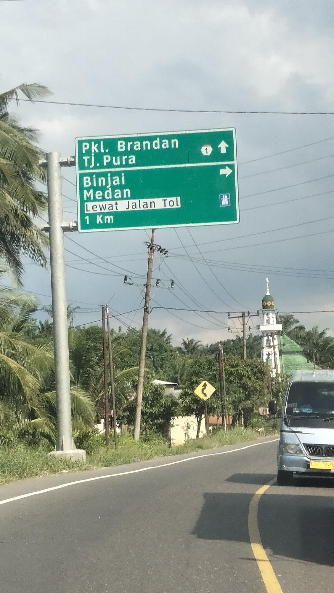 The image shows a green road sign with white text in Indonesian. The sign indicates directions to "Pkl. Brandan Tj. Pura" with a route number "1" and an upward arrow. Below that, it directs to "Binjai Medan" with a right arrow, and then "Lewat Jalan Tol 1 Km" (Via Toll Road 1 Km) with a symbol indicating an entrance to a toll road. The sign is supported by a metal pole on the left and a wooden utility pole on the right, which has multiple wires running across it.

In the background, there are palm trees on the left, and further back, a mosque with a green dome and a white minaret is visible. A silver van is driving on the asphalt road in the foreground on the right. The road has a white line on the left edge and a yellow curved line in the center. The sky is cloudy. The location is identified as Tangkahan Durian, Indonesia.