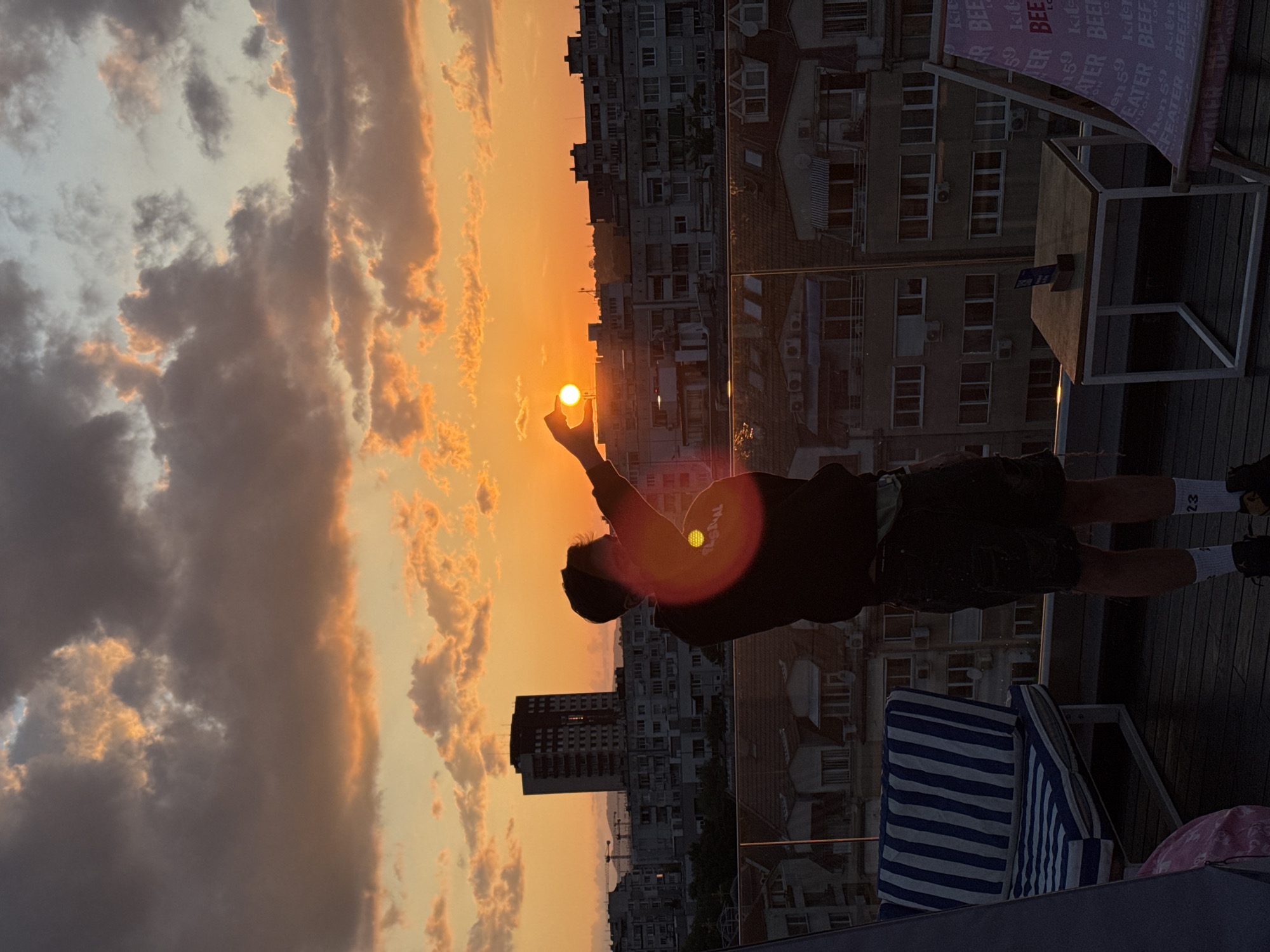 The image depicts a rooftop scene at sunset in Sofia, Bulgaria. The sky transitions from a deep orange and yellow near the horizon to a lighter blue-grey at the top, interspersed with scattered dark clouds that are illuminated orange and pink from below. The sun, a bright yellow-orange orb, is positioned low on the horizon, creating a prominent circular lens flare that illuminates the central figure.
A silhouetted individual, appearing to be an adult male, stands on a dark wooden deck with their back mostly to the viewer, facing the setting sun. They are wearing a dark long-sleeved top, dark shorts, white socks (one displaying "23"), and dark sneakers. A light-colored fanny pack is visible around their waist. The person's right arm is extended upwards, with their hand positioned to create the optical illusion of pinching or holding the sun.
The rooftop area features a transparent glass railing separating it from the cityscape. To the left, a blue and white striped cushion, part of outdoor seating, is visible. To the right, a small square wooden table with white metal legs holds a dark, upright object. Further right, a pink and white striped fabric lounge chair, with repeated "BEER" text, is partially visible.
Beyond the railing, a dense urban landscape unfolds under the sunset glow. Numerous multi-story residential and commercial buildings, predominantly in muted greys and silhouettes, stretch across the mid-ground. A prominent, taller, dark-colored high-rise building is visible to the left of center. Distant construction cranes and the faint outline of hills or mountains are discernible on the horizon. The foreground elements are largely in deep shadow, contrasting sharply with the bright, warm light of the setting sun.