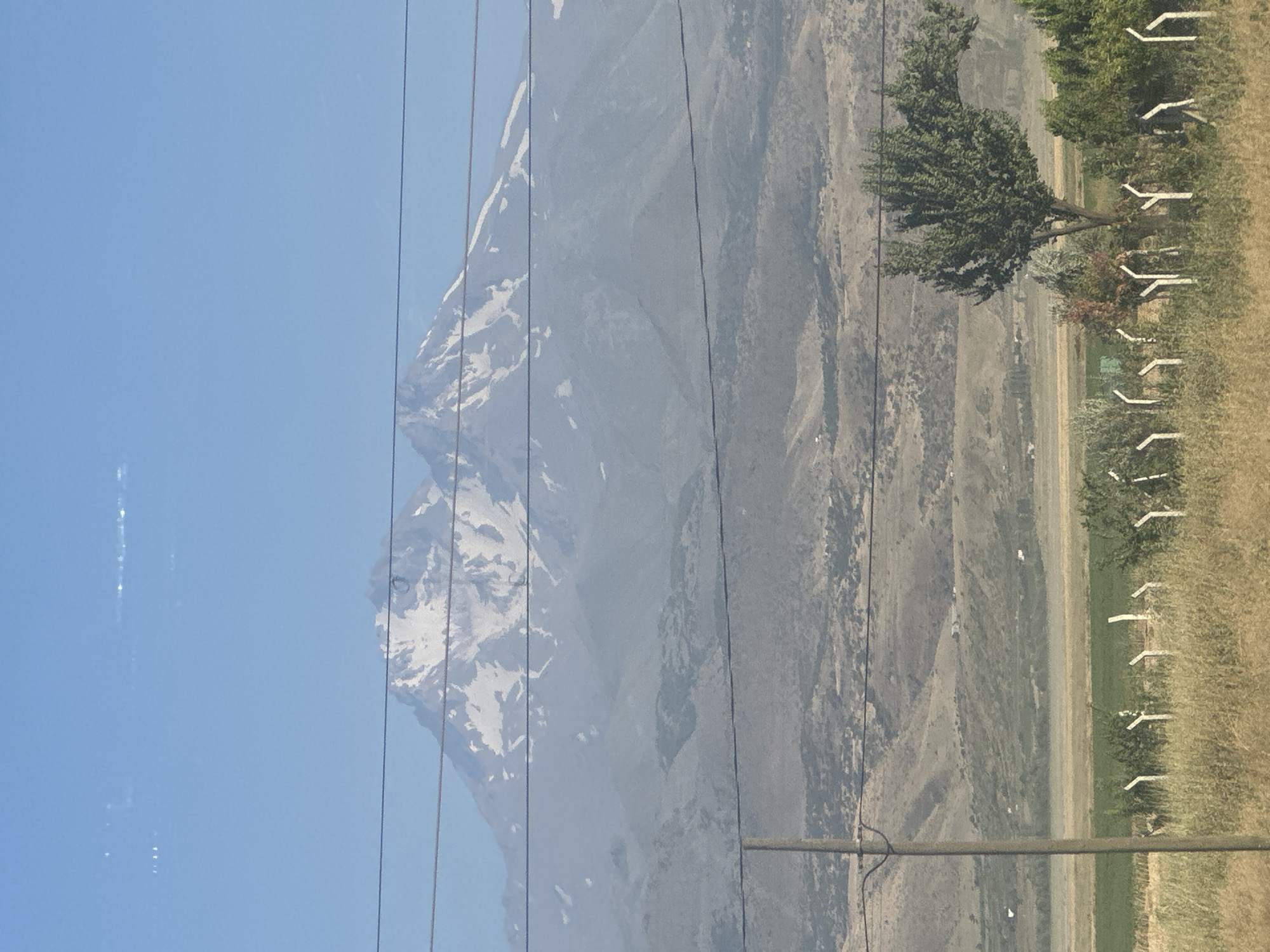 The image is a landscape photograph taken in the city of İncesu, Turkey, depicting a view of a snow-capped mountain in the distance. The mountain has a rugged texture and is partially obscured by haze. In the foreground, there is a dry, grassy field with a few sparse trees and a fence made of white posts. Power lines run horizontally across the frame, obscuring parts of the mountain and sky. The sky is a clear blue with some faint streaks of light that might be lens flare or atmospheric phenomena. The overall scene suggests a rural or semi-rural environment with a prominent natural landmark.