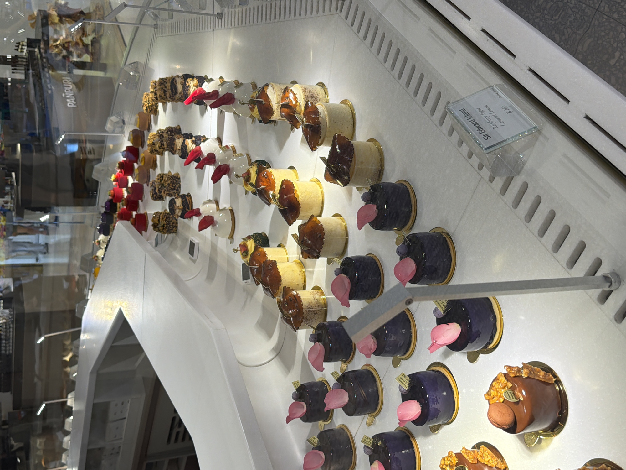 This is a photograph of a pastry display case, likely taken in a bakery or dessert shop. The brand “PANPURI” is visible, which is a luxury lifestyle brand based in Bangkok, Thailand. The display case contains numerous small, round pastries. These appear to be arranged in rows, with different flavors and toppings, including dark-glazed pastries with pink flower petal decorations, light-colored pastries with brown leaf-shaped toppings, and chocolate pastries. One pastry has a small macaron on top. A glass sign is visible, which shows "Sir Edward Island Raspberry Figue Caramel Lemon $2.65". The lighting in the display case is bright, highlighting the pastries.