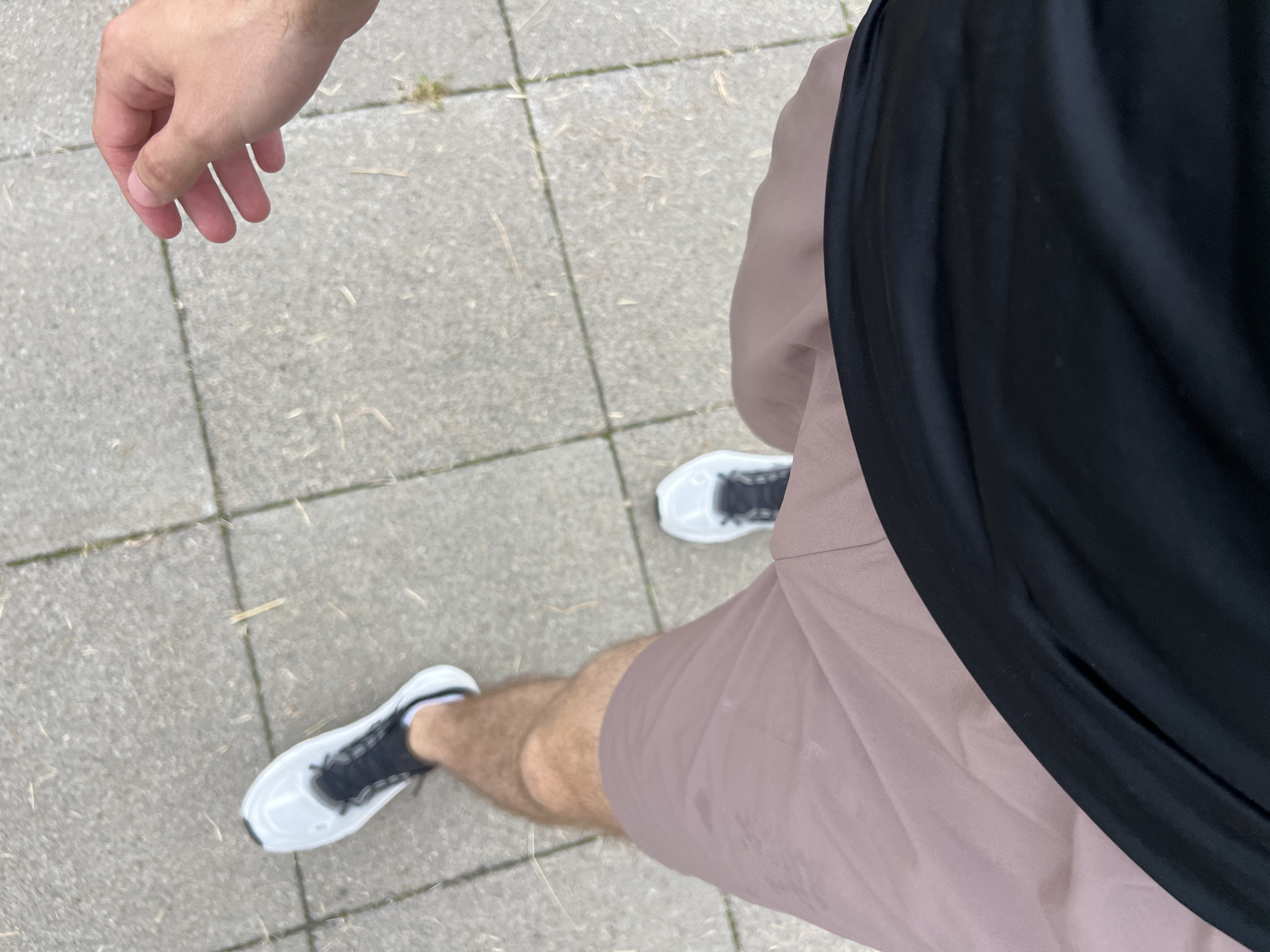 The image shows a person's legs and hand from a first-person perspective, as they walk on a paved sidewalk. The person is wearing light-colored sneakers with black laces, light brown shorts, and a black t-shirt. A hand is visible in the upper right corner, with fingers slightly curled. The ground is made of square concrete tiles with some dry grass and debris between them. There are no discernible landmarks or features that would indicate the location within the city of Krasnoyarsk, Russia.