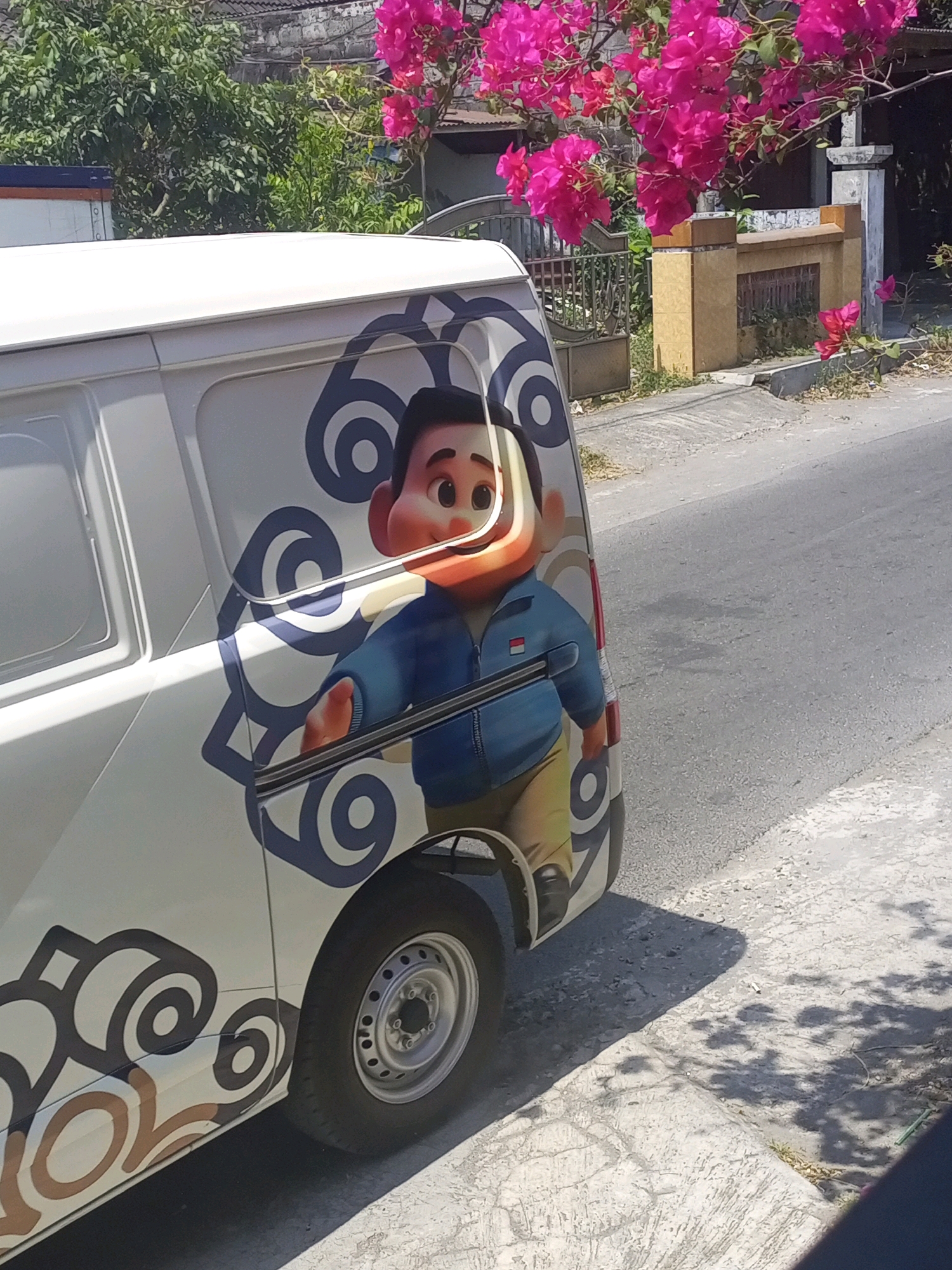 In Ngasem, Indonesia, a white van is parked on the side of a paved road. The van is decorated with a cartoon character and various swirl designs. The character appears to be a young boy with large, curious eyes, wearing a blue jacket and yellow pants. Behind the van, a stone wall with ornate pillars and a metal fence can be seen. Bright pink bougainvillea flowers are in full bloom, adding a splash of color to the scene. The ground next to the road is a mix of concrete and dirt, with some dry grass and shadows cast by trees.