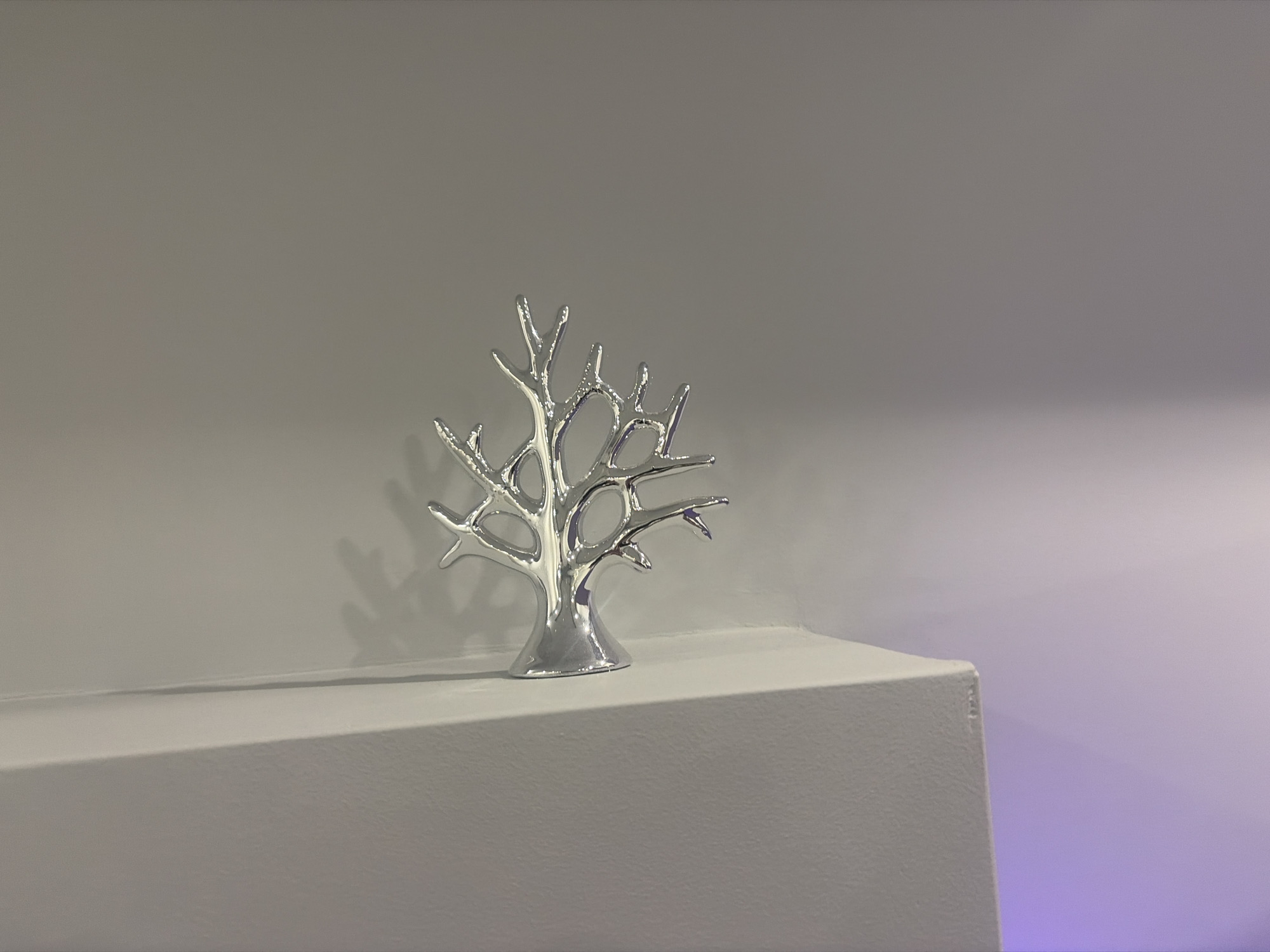 A silver decorative tree sculpture with an open, branching design is placed on the edge of a white, textured surface. The surface appears to be a shelf or ledge, with a subtle gradient of light on the wall behind it, shifting from a neutral gray to a soft purple glow. The sculpture's reflective surface catches the light, creating highlights and shadows. The background is a plain, neutral gray wall. This scene is located in a domestic indoor setting in Podgumer, Bulgaria. There are no people or animals present in the image.