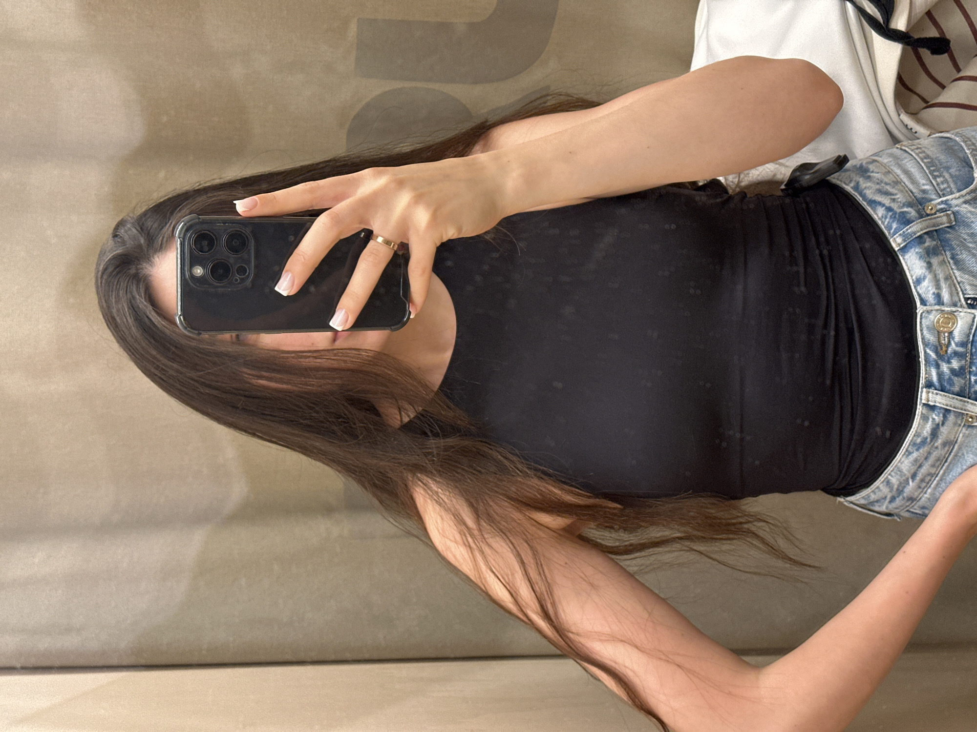 A woman is taking a selfie in a fitting room, likely in a clothing store, in Kazan, Russia. She is wearing a black top and light blue jeans, with a white and striped garment draped over her shoulder. She is holding a black smartphone, obscuring her face, with her left hand adorned with a gold ring. The background consists of a neutral-colored wall.