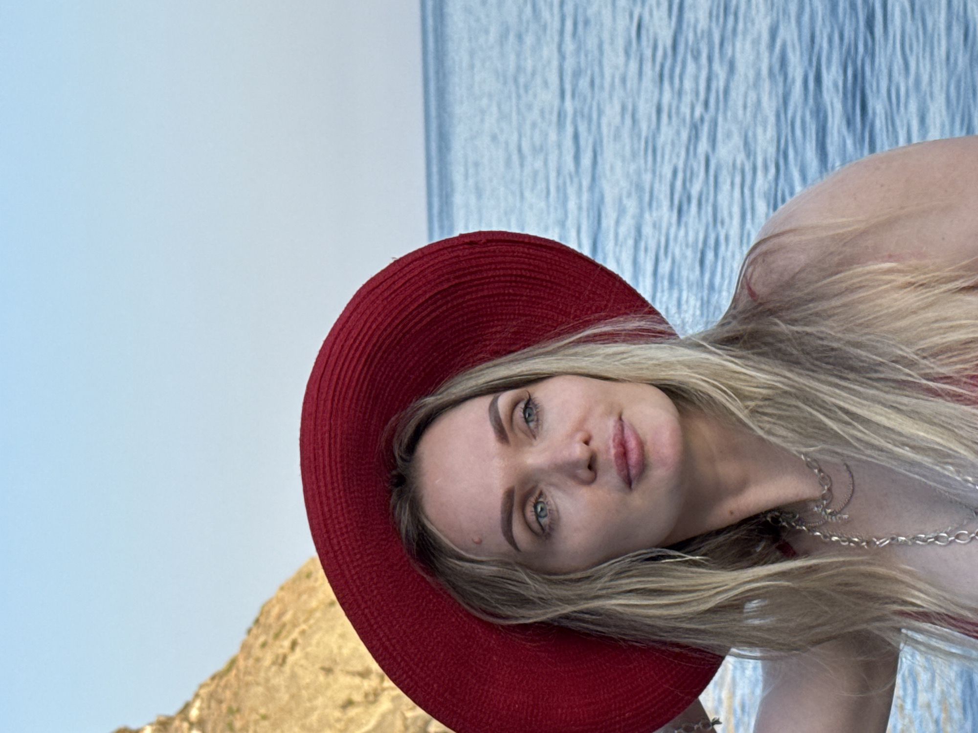 A young woman with blonde hair and blue eyes is captured in a close-up portrait, wearing a large red hat. She is positioned outdoors, with a body of water and a rocky cliff face visible in the background. The sky is a clear blue. The woman is looking off to the right, with a thoughtful expression. She is wearing a red top and a silver necklace. The scene suggests a coastal location, possibly near the city of Utios, Ukraine.