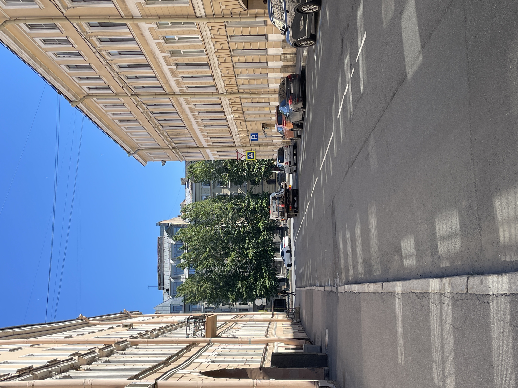 This is a ground-level, outdoor shot taken on a street in Saint Petersburg, Russia, on a clear, sunny day, likely in the afternoon due to the long shadows cast by the buildings and trees. The image is oriented vertically, with the top of the frame showing a bright blue sky and the bottom showing the pavement and a building.
The scene depicts a street lined with buildings on both sides. The building on the left is a multi-story structure with a light-colored facade and numerous windows. The building on the right appears to be a taller, more modern building with a grey facade, partially obscured by a large, lush green tree. Several cars are parked along the right side of the street, including a white van at the far end and a black SUV closer to the foreground. A parking sign is visible, indicating a parking area.
The pavement is asphalt with white markings. The sunlight creates distinct patterns of light and shadow on the road surface, suggesting a bright, sunny day. There are no people prominently featured in the image, though a person can be faintly seen walking on the sidewalk in the distance. The overall mood is calm and everyday, capturing a typical urban street scene. No legible text is present in the image.