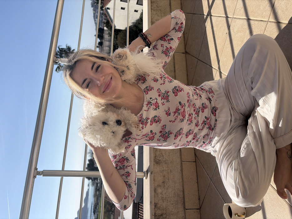 A woman with blonde hair, wearing a floral shirt and white pants, sits cross-legged on a balcony. She's holding two small white dogs close to her neck. A coffee cup sits on the ground near her feet. The balcony railing is visible, with a cityscape and blue sky as the backdrop. The sun is shining, casting shadows on the tiled floor.