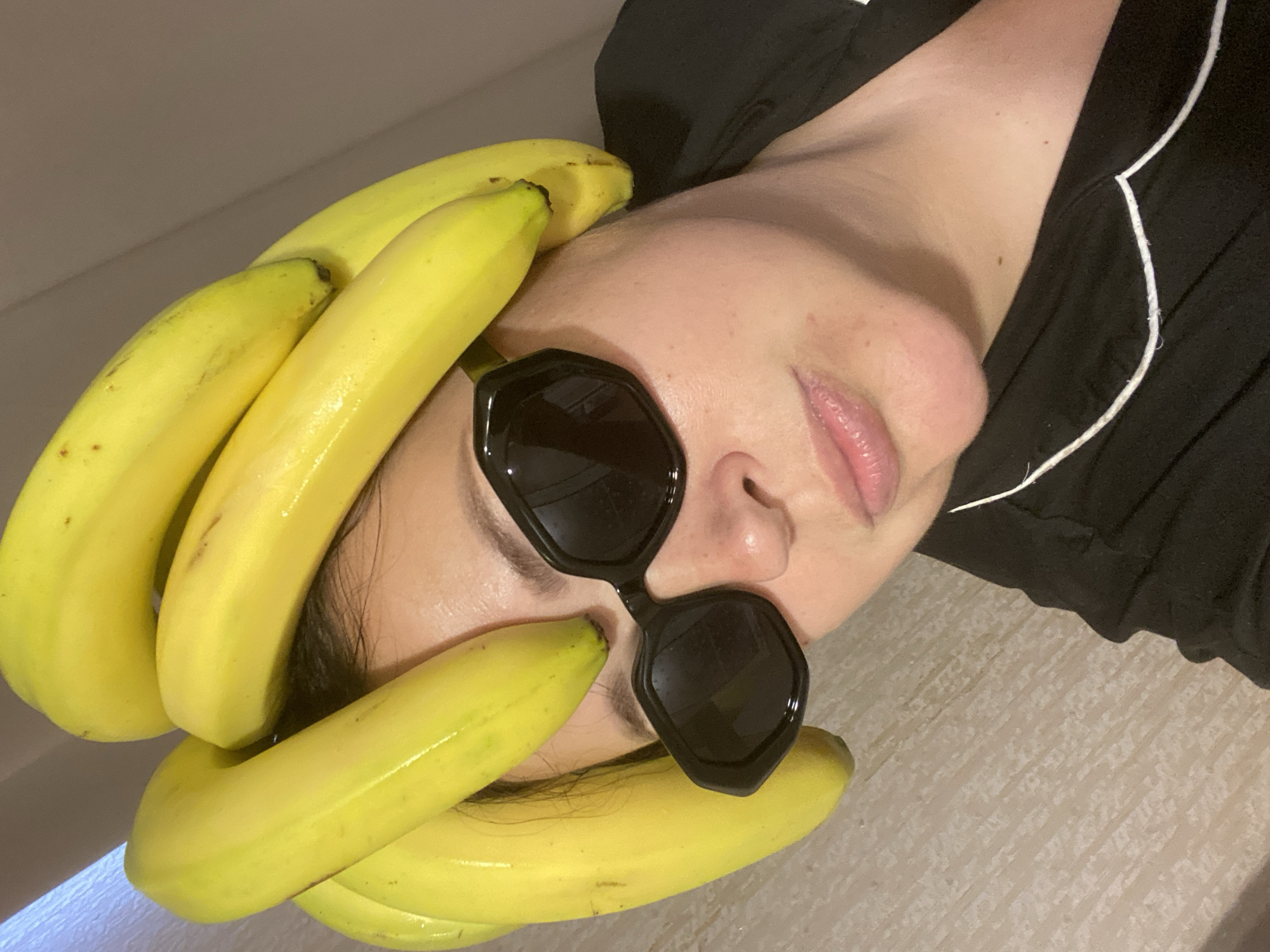 A close-up shot features a person lying down with several bananas arranged around their head, partially obscuring their forehead and face. The person is wearing sunglasses. They are wearing a black top with white piping along the collar. The background appears to be a light-colored surface. The image is shot at an angle, giving an off-kilter perspective. It is not possible to determine the location of the image from the image itself, including whether or not it is in the city of Sochi, Russia.