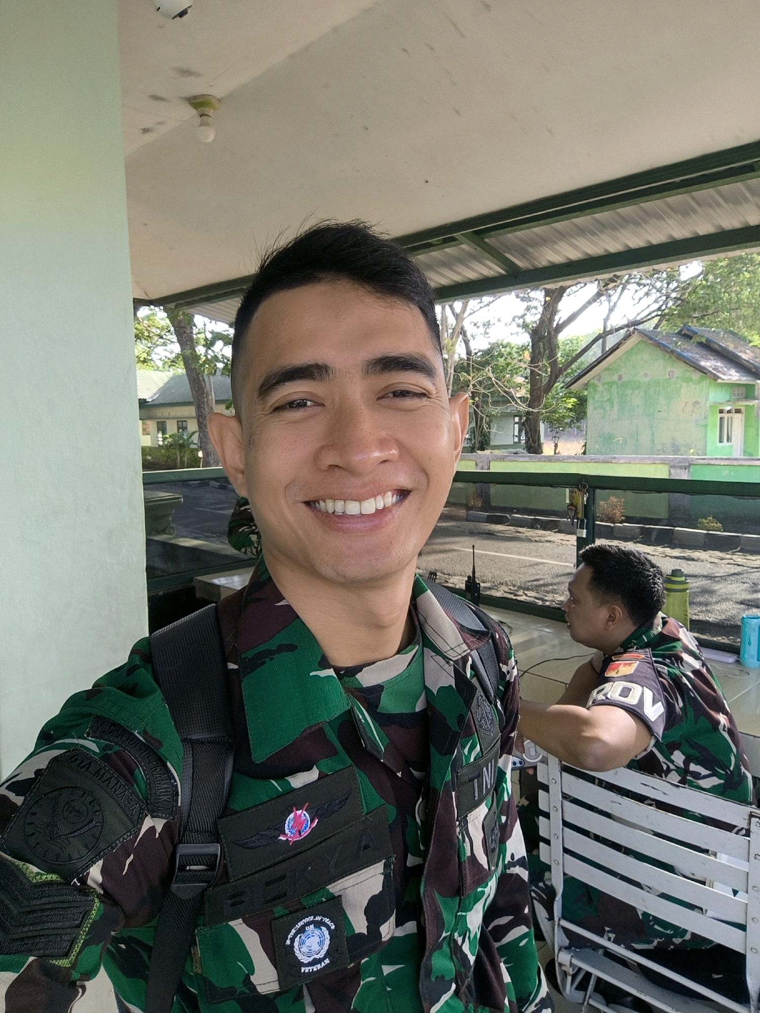 A medium close-up selfie shows a smiling man in a camouflage uniform with a black backpack. He is standing in front of a light green wall and a covered walkway. In the background, another man in a similar uniform is seated at a white table. Behind him, a light green building and trees are visible. The overall setting appears to be an outdoor military facility.