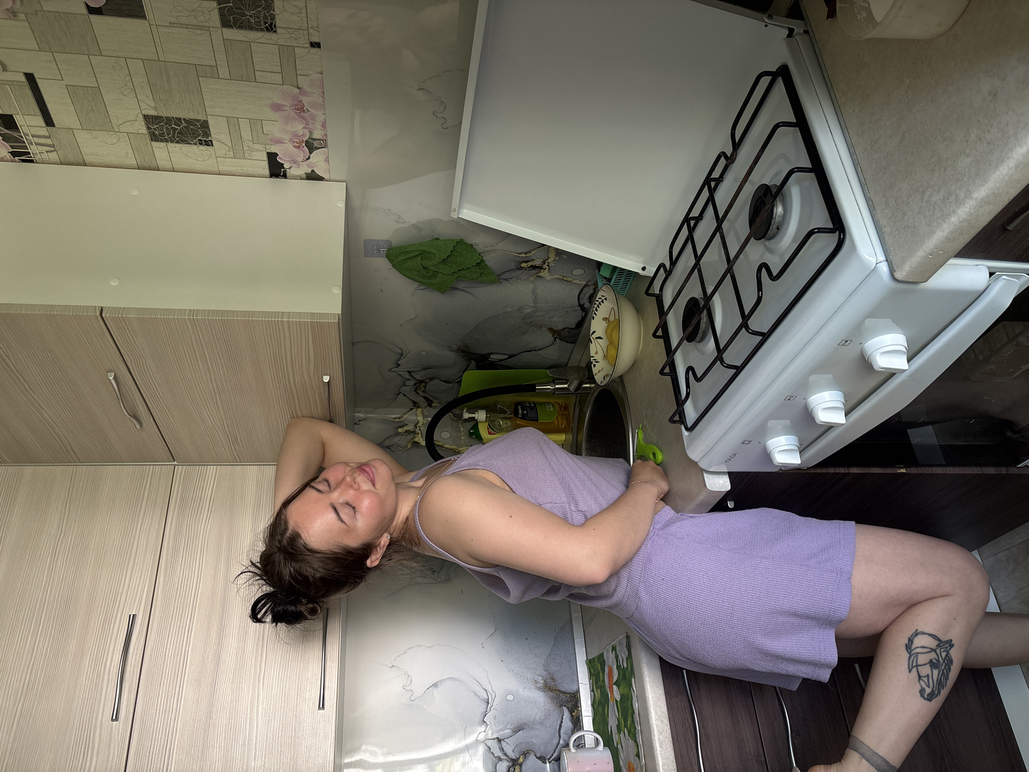 A woman lies in a kitchen in Izhevsk, Russia. She has dark hair pulled into a bun and wears a light purple dress. Her eyes are closed. She is lying on her side near a kitchen sink, gas stove, and cabinets. A tattoo of a horse's head is visible on her left leg. The kitchen has patterned wallpaper and various kitchen items near the sink.