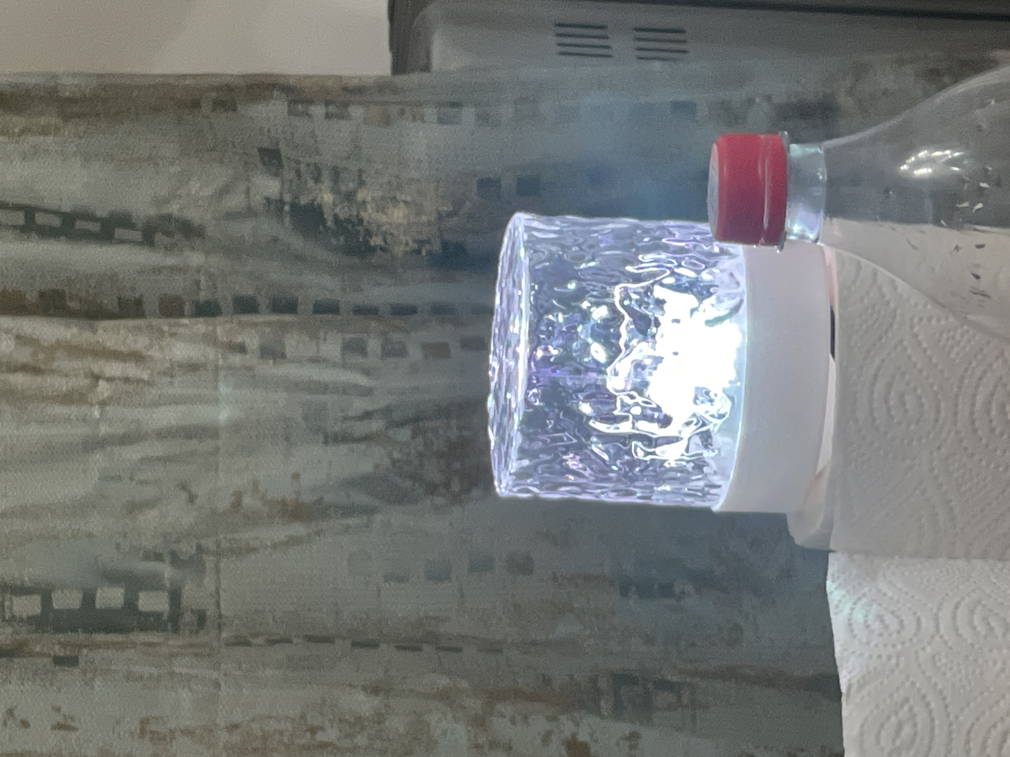 A close-up, eye-level, indoor shot features a clear, textured cylindrical lamp emitting a bright white and purple light in the center of the frame. The lamp sits atop a white base, which is positioned on a roll of white paper towels to the left. To the right of the lamp is the top portion of a clear plastic bottle filled with a clear liquid and capped with a red plastic lid. The background consists of a grey, textured curtain with a subtle checkered pattern. The overall lighting is dim, with the lamp being the primary light source. There is no indication of the city of Bishkek City, Kyrgyzstan.
