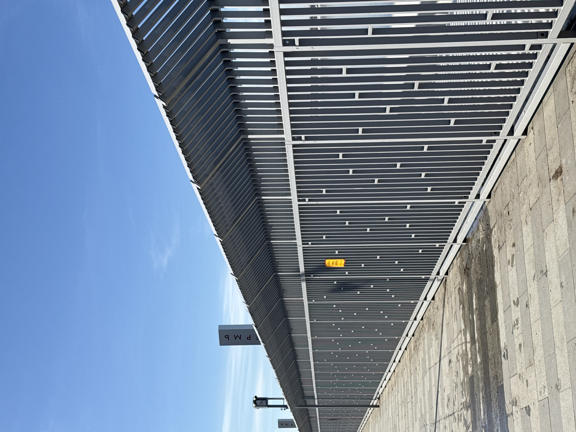 The image displays a section of the Perm River Embankment along the Kama River in Perm, Russia. The primary subject is a long, modern grey metal barrier extending diagonally from the lower right to the mid-left of the frame, under a clear blue sky. This barrier is constructed from numerous vertical rectangular slats, some solid and some featuring smaller rectangular cutouts, forming a repetitive pattern. A slatted metal canopy, angled upwards, extends from the top edge of the vertical barrier. A small, rectangular yellow sign with dark, illegible text is affixed to the barrier in the central-right portion of the frame.
Adjacent to the barrier, a paved walkway made of light-grey rectangular tiles shows visible patches of dampness or discoloration. In the background, behind the barrier, two tall, grey columnar signs are present. The most prominent sign clearly displays the Cyrillic letters "Р М Ь" stacked vertically. A second, similar columnar structure is partially visible further to the left. No individuals or active subjects are present in the scene. The environment suggests a public urban space, likely a promenade or pedestrian area, on a sunny day.