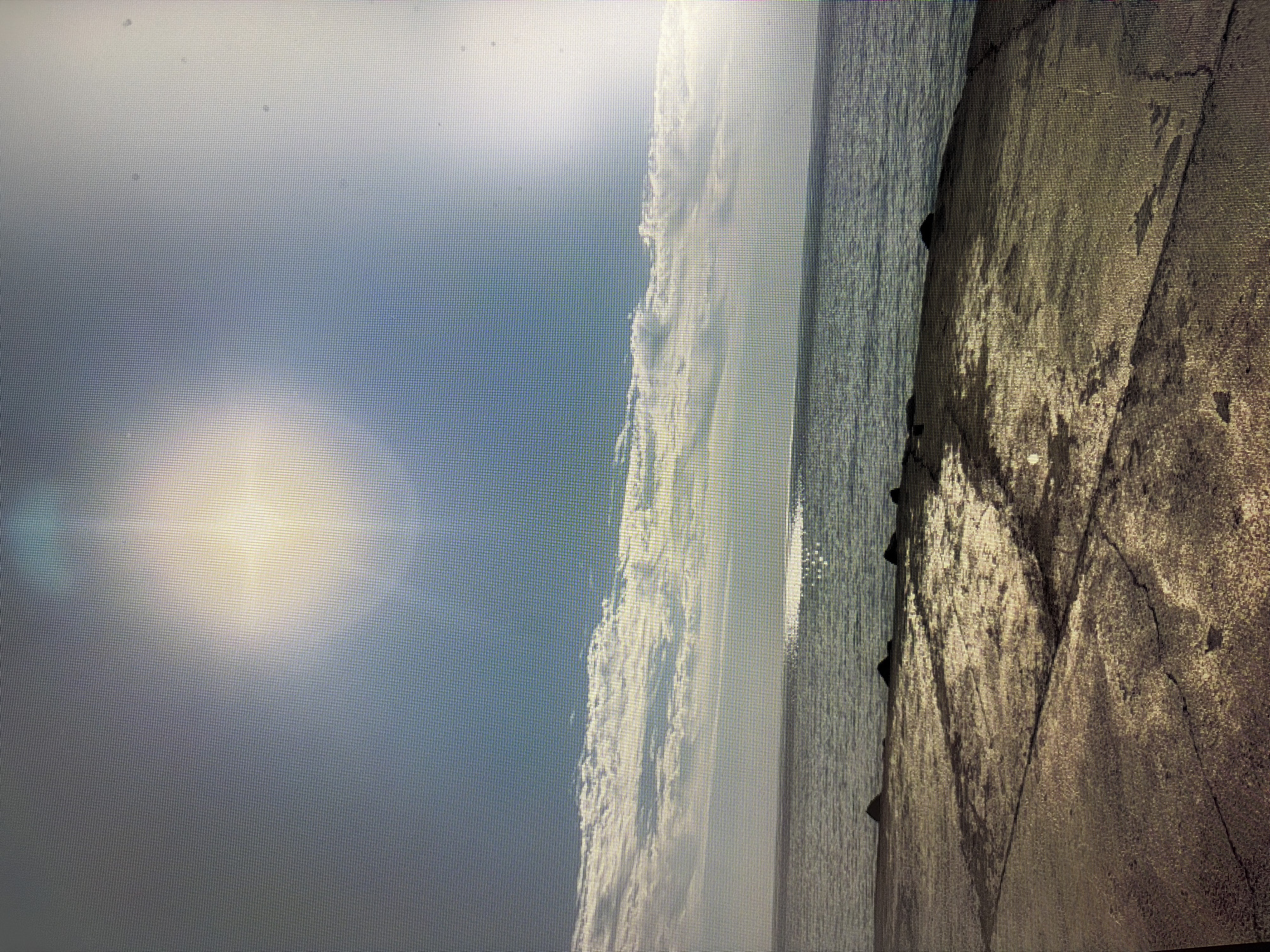 The image depicts a wide, static shot of a body of water meeting a concrete shoreline under a bright, partly cloudy sky. The sun is high, casting a bright glare on the water's surface and creating a hazy effect in the sky. The foreground is dominated by weathered, cracked concrete, suggesting an industrial or public access area. Dark, irregularly shaped rocks are visible along the edge of the water. The water itself appears calm, with subtle ripples reflecting the sky. The clouds are arranged horizontally, with a thin band of white clouds stretching across the horizon, contrasting with the clear blue sky above. There are no people or visible signs of activity in the scene. The location is specified as Mirnoye, Ukraine.