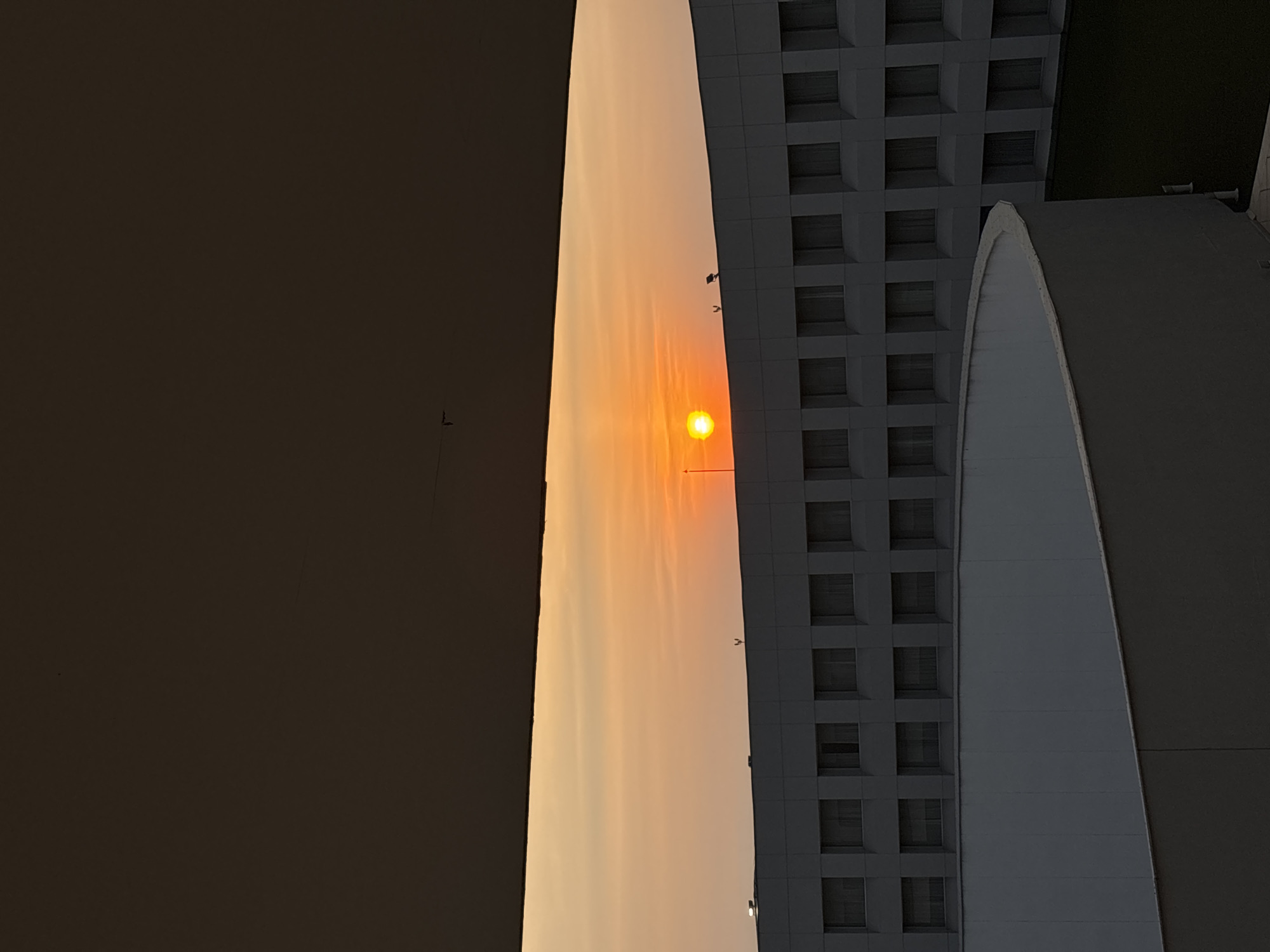 The image depicts a sunset view between two buildings, one with a gridded facade and the other appearing more monolithic, likely taken from an elevated position within Mexico City, Mexico. The setting sun is partially obscured by haze, casting a warm orange glow across the sky. Utility poles or other vertical structures are visible in the distance.