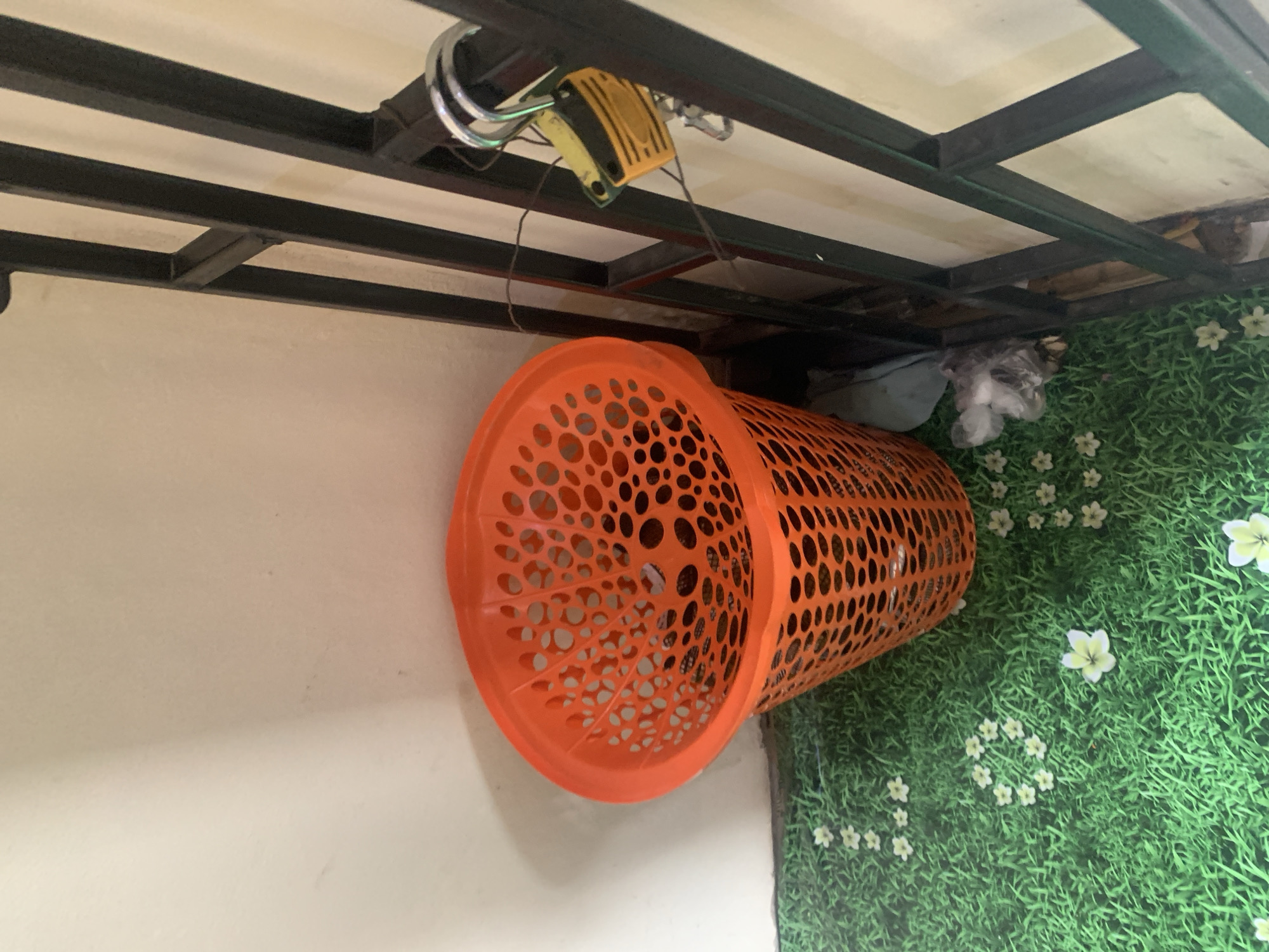 An orange plastic laundry basket sits on a green floor with a grass and flower pattern. To the right of the basket is a black metal grate with a padlock and what appears to be a yellow tag attached to it. Behind the basket and grate is a light-colored wall. The scene is indoors, likely in a room.