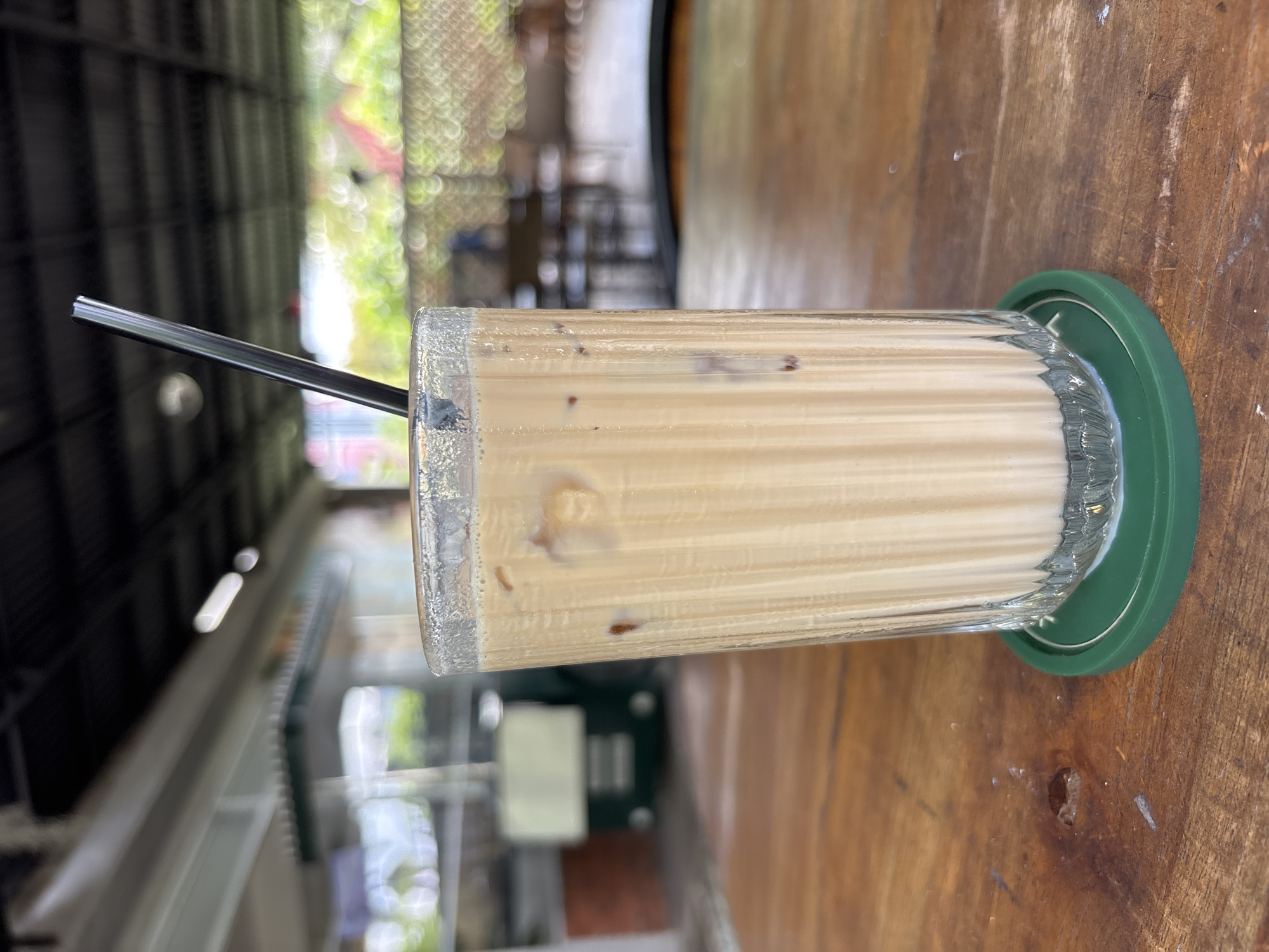A close-up shot of a beverage, likely iced coffee or a similar drink, in a clear, ridged glass with a black straw. The glass is placed on a green coaster on a wooden table. The drink has a light brown color with visible ice cubes or particles. The background shows a blurred outdoor environment, possibly a cafe or restaurant. The setting appears to be Kuta Selatan, Indonesia, based on the architectural style and tropical vegetation visible through the blurred background. No people are visible in the foreground or immediate background.