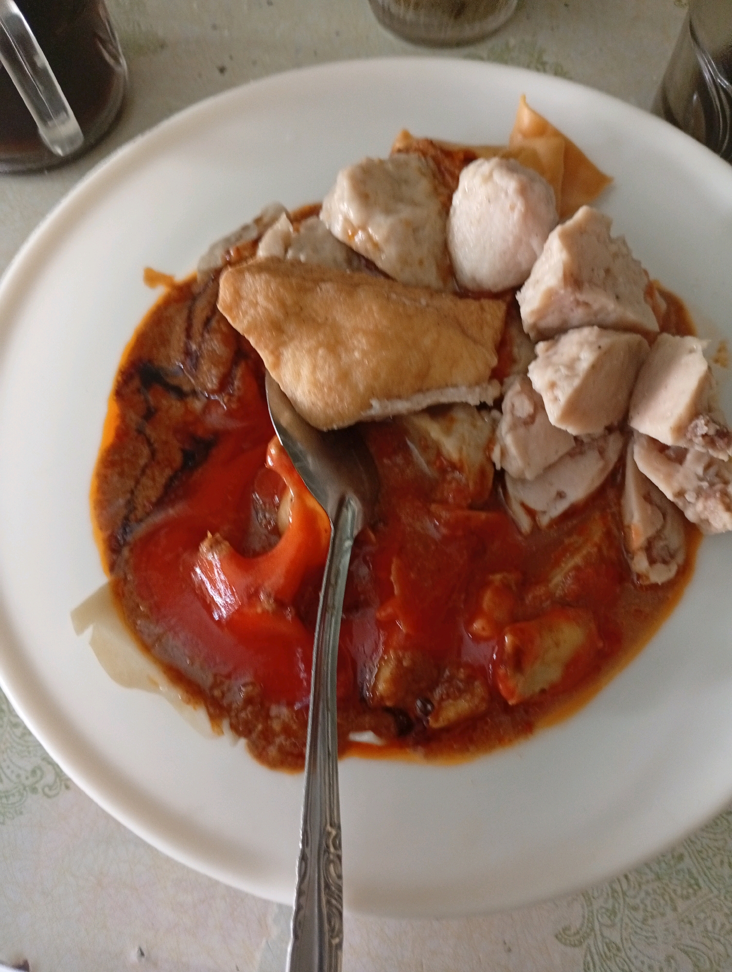 A close-up, overhead shot captures a white plate filled with a dish of food, likely served at a restaurant in East Jakarta, Indonesia. The plate contains several pieces of meatball-like items, some fried tofu, and a thick, reddish-brown sauce with visible chunks of what appears to be chili. A silver spoon rests on the plate, partially submerged in the sauce. In the upper left corner, a glass container with a dark liquid and a straw is partially visible. The table surface beneath the plate has a light-colored, patterned texture. The lighting is soft and natural, creating minimal shadows.