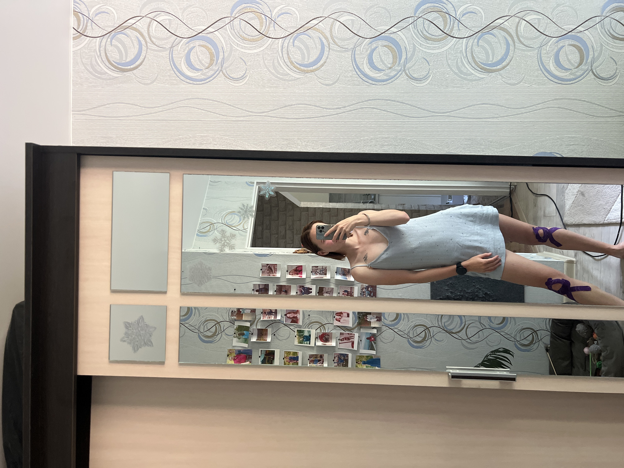 The image depicts a young woman taking a mirror selfie in what appears to be a bedroom or dressing area. She is wearing a light blue slip dress and has purple kinesiology tape applied to her knees. In the background, a large wardrobe with sliding doors is visible. One door is a mirror reflecting the woman and the room behind her, which is decorated with wallpaper featuring a swirling pattern of circles and lines. A section of the wardrobe door is a pale pink, likely a painted panel. On the wardrobe door, above the mirror, are two square decorative panels; one with a silver snowflake design and another plain rectangular panel. Inside the mirrored section of the wardrobe, several small photos are attached to the wall, creating a collage. The floor is partially visible, showing a rug. The overall setting suggests an indoor domestic environment. The woman is holding a smartphone in her right hand, angled to capture the reflection. Her left hand is resting on her hip. The lighting appears to be natural, coming from an unseen source.
