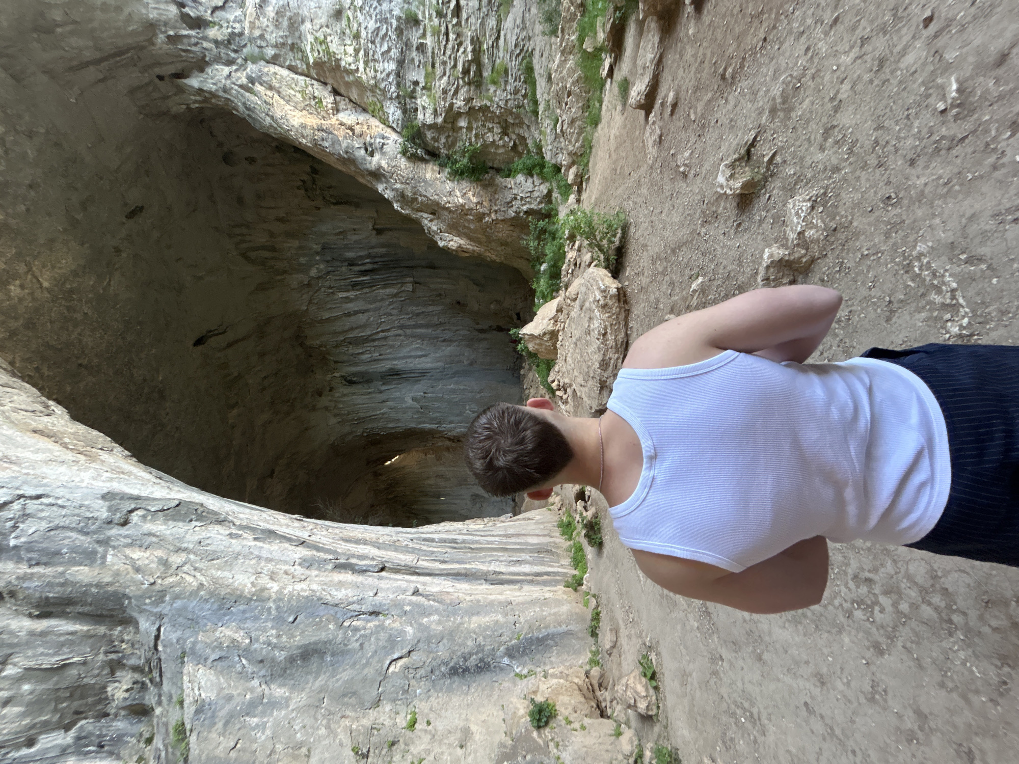 A man in a white tank top stands at the edge of a large cave, looking down into it. The cave has rocky walls with horizontal striations. The ground surrounding the cave entrance is a mix of dirt and rocks. Some greenery clings to the rock face near the entrance. The man appears to be exploring the Devetashka cave located near Karlukovo, Bulgaria.