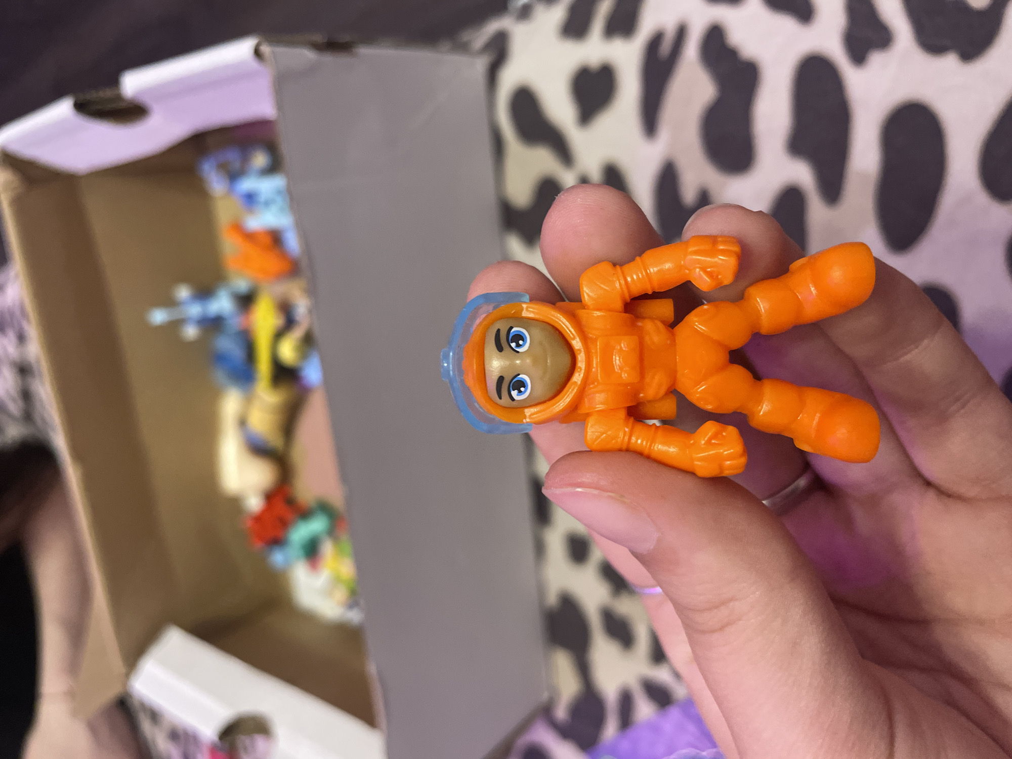 A close-up shot captures a small, orange astronaut figurine being held between a person's thumb and index finger. The figurine has a blue helmet with a clear visor, and its face is painted with blue eyes and a light brown skin tone. The astronaut's suit features details like a chest plate, arm joints, and articulated legs. In the blurred background, a cardboard box is visible, containing various other small toys and objects, some of which appear to be colorful figurines and building blocks. The patterned surface beneath the box and the fingers suggests a textile, possibly a blanket or clothing, with a cow-print or similar abstract design. The lighting is dim, casting soft shadows and highlighting the textures of the figurine and the skin. The scene is devoid of any recognizable landmarks or elements that would indicate a specific location within Ladozhskii Trudposelok, Russia.
