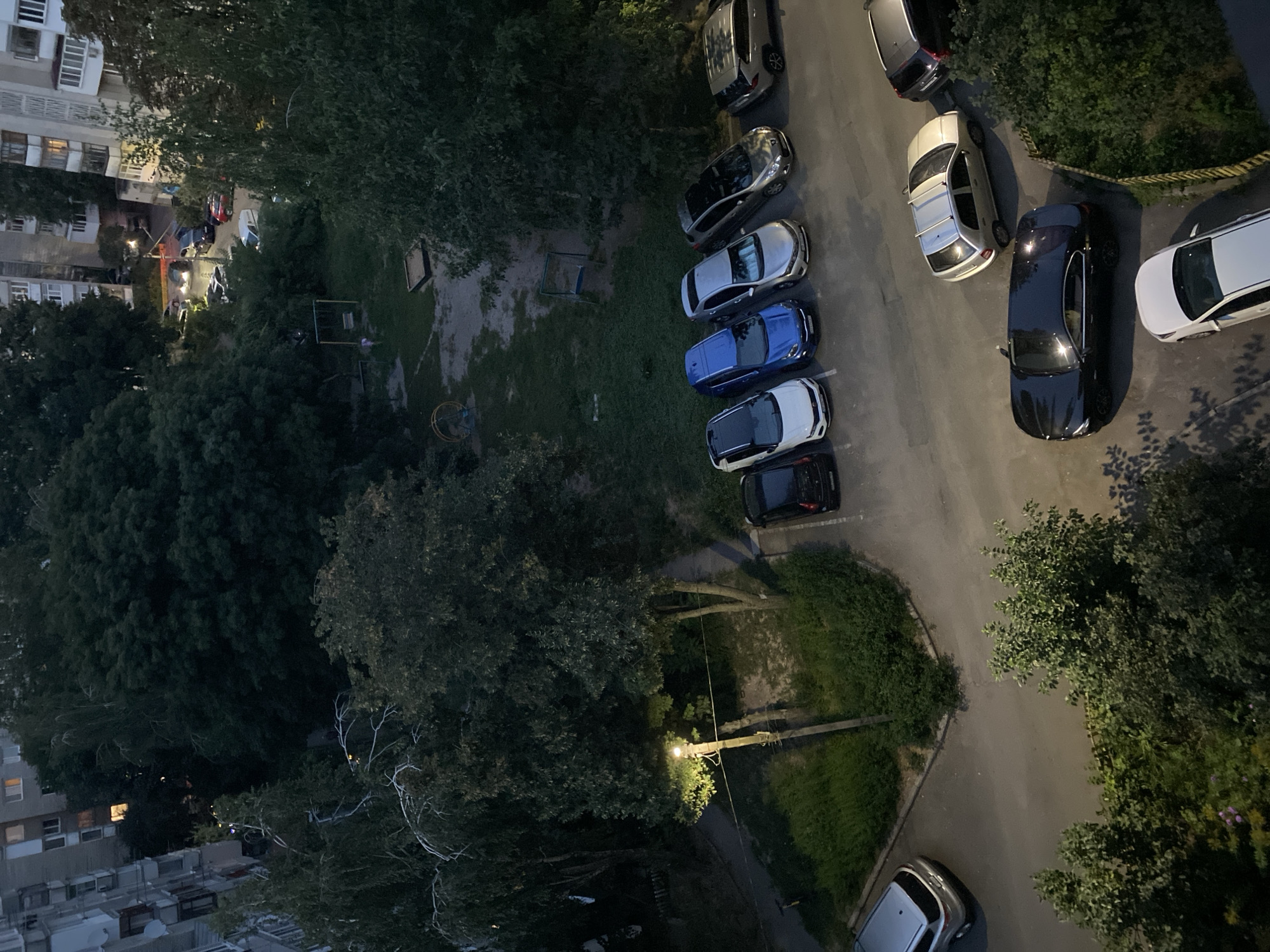 An outdoor, medium-high angle, eye-level shot captures a residential courtyard in Vinnytsia, Ukraine, in what appears to be twilight or early evening. The scene is dominated by mature trees with dense foliage, casting shadows across a paved area where several cars are parked. To the left, a multi-story apartment building, typical of Soviet-era architecture, features visible windows, some illuminated. A street lamp stands prominently near the center-left of the frame, casting a warm glow on the surrounding trees and pavement.
In the foreground, the paved area shows various cars parked in designated spots. From left to right, these include a silver SUV, a black sedan, and a white sedan. Further into the courtyard, a row of cars is parked: a black sedan, a white hatchback, a blue hatchback, a silver hatchback, and a gray hatchback. A silver SUV is parked at the far right of this row. On the right side of the paved area, a dark-colored SUV is parked near a dense green hedge.
The background reveals a small, grassy playground area with a swing set and a sandpit. The overall impression is a typical urban residential setting, suggesting a quiet evening in a community. There are no visible people in the frame.
