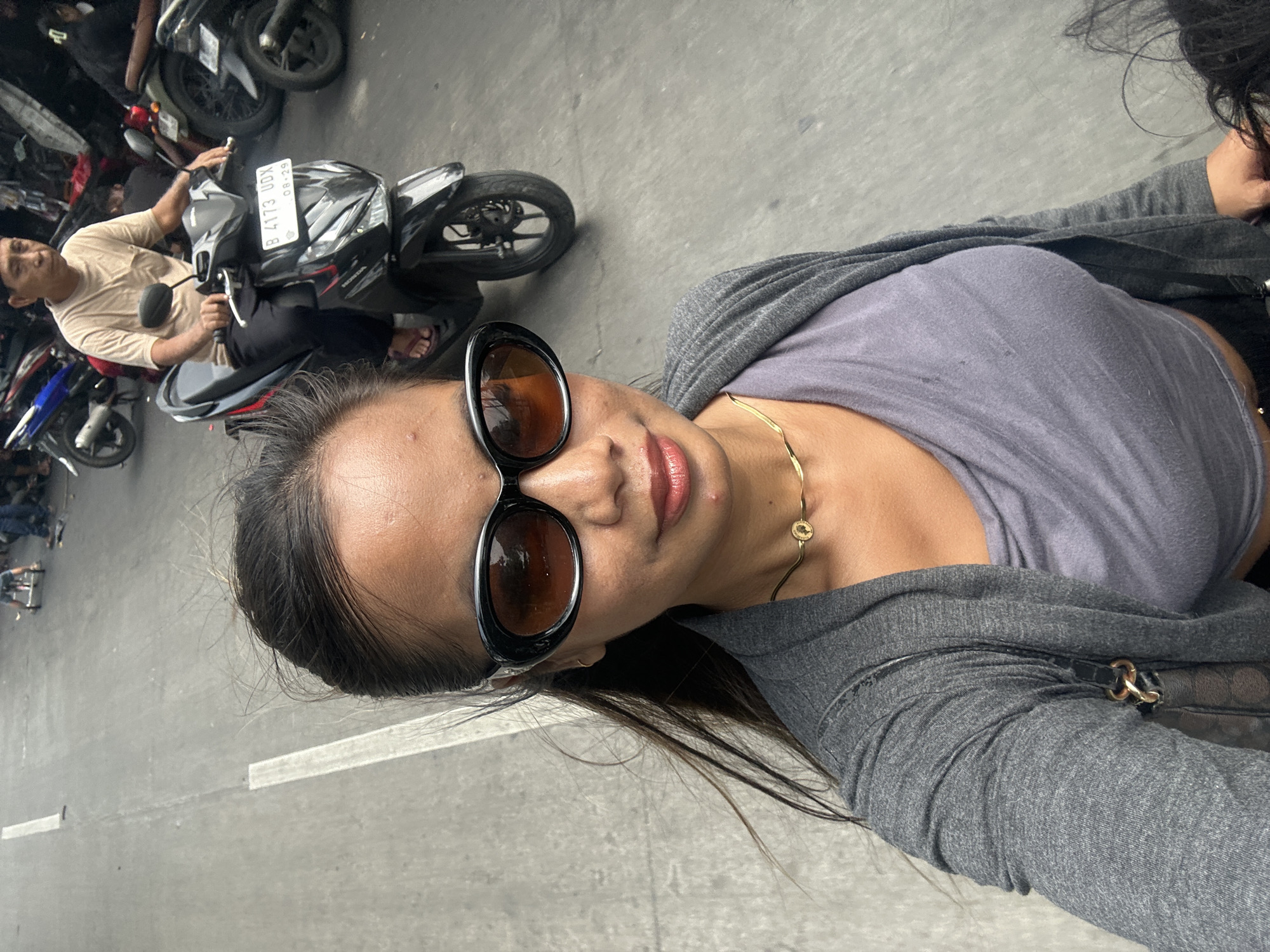 The image shows a woman in sunglasses and a gray top and jacket. In the background, a man is sitting on a motorcycle with a license plate reading "B 4173 UDX." Several other motorcycles are parked nearby. The scene appears to be a street in North Jakarta, Indonesia.