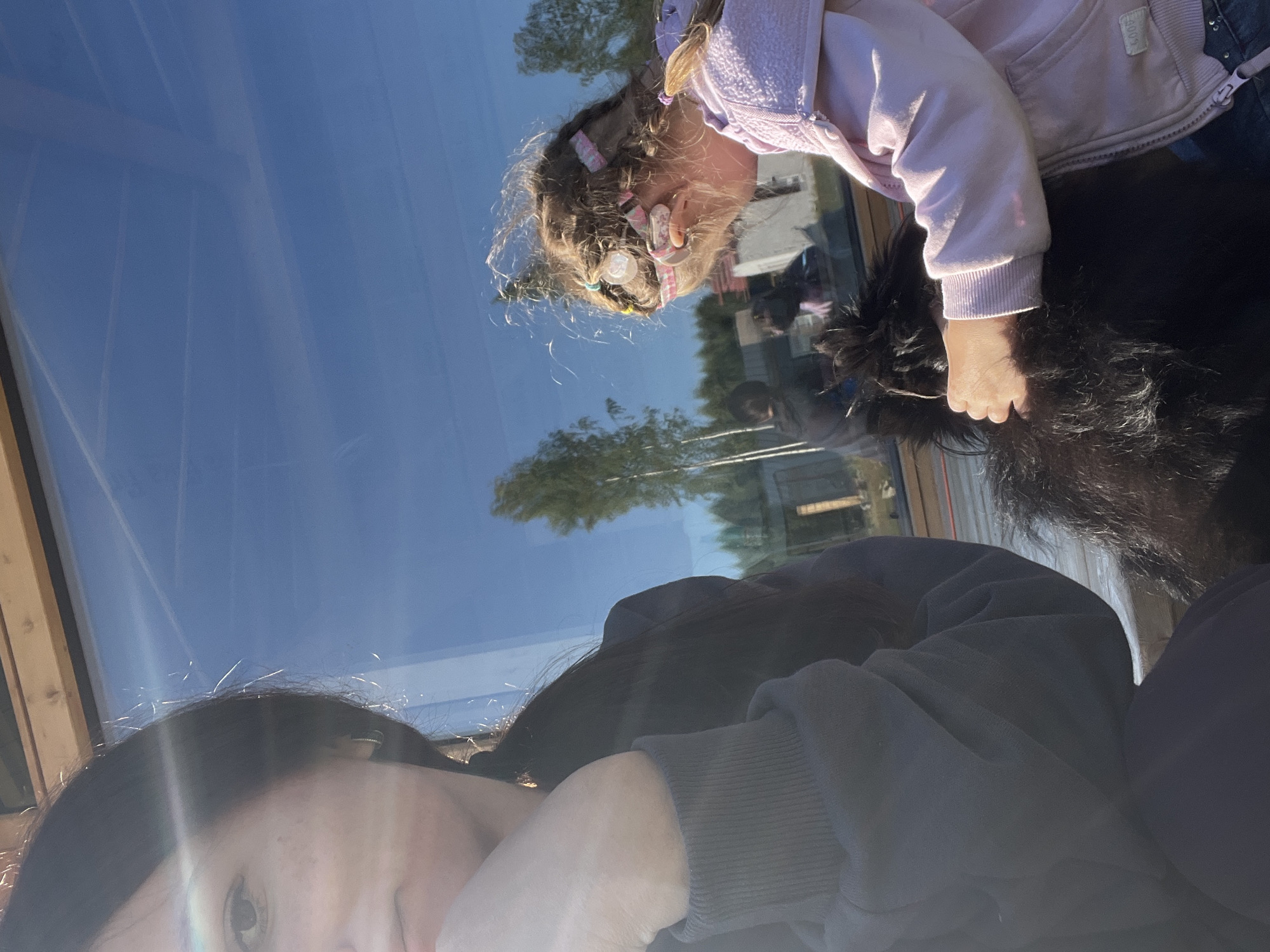 In Imeni Morozova, Russia, a young girl with braided hair, wearing a pink hoodie and blue jeans, pets a fluffy black dog. She is reflected in a large glass window, which also shows the blue sky and trees. To the left, a woman with dark hair is partially visible, her face turned away from the camera and her hand resting on her chin. The scene appears to be outdoors, possibly in a backyard or near a building with a large glass facade. The sunlight casts long shadows and creates a bright, clear atmosphere.