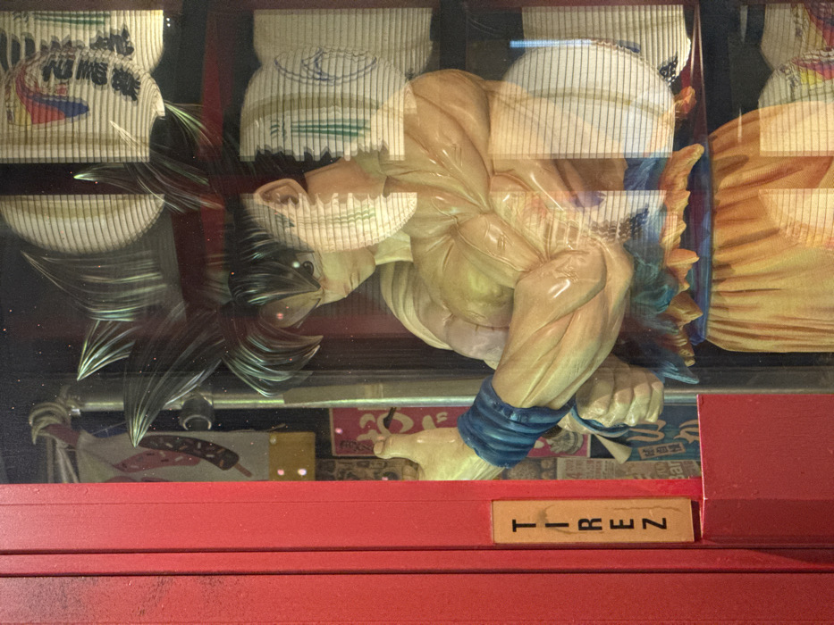 The image displays a detailed statue of the anime character Goku from Dragon Ball, presented in his Super Saiyan form with spiky black hair, wearing his traditional orange gi with blue wristbands and belt. The statue, showcasing a muscular physique and a determined expression, is situated inside a reflective glass display case. Behind Goku, the interior of the case features a dark, cluttered background adorned with various Japanese cultural elements and posters, including one depicting what appears to be Dango skewers. To the immediate left of the display, a prominent bright red vertical doorframe or panel is visible. Affixed to this panel is a rectangular, light-colored sign with the French word "TIREZ" (PULL) inscribed vertically in black capital letters, indicating the scene is located in Paris, France. Significant reflections on the glass surface superimpose a pattern of horizontal blinds and faint images of Japanese paper lanterns (chōchin) with black calligraphy, partially obscuring the statue's face and body.