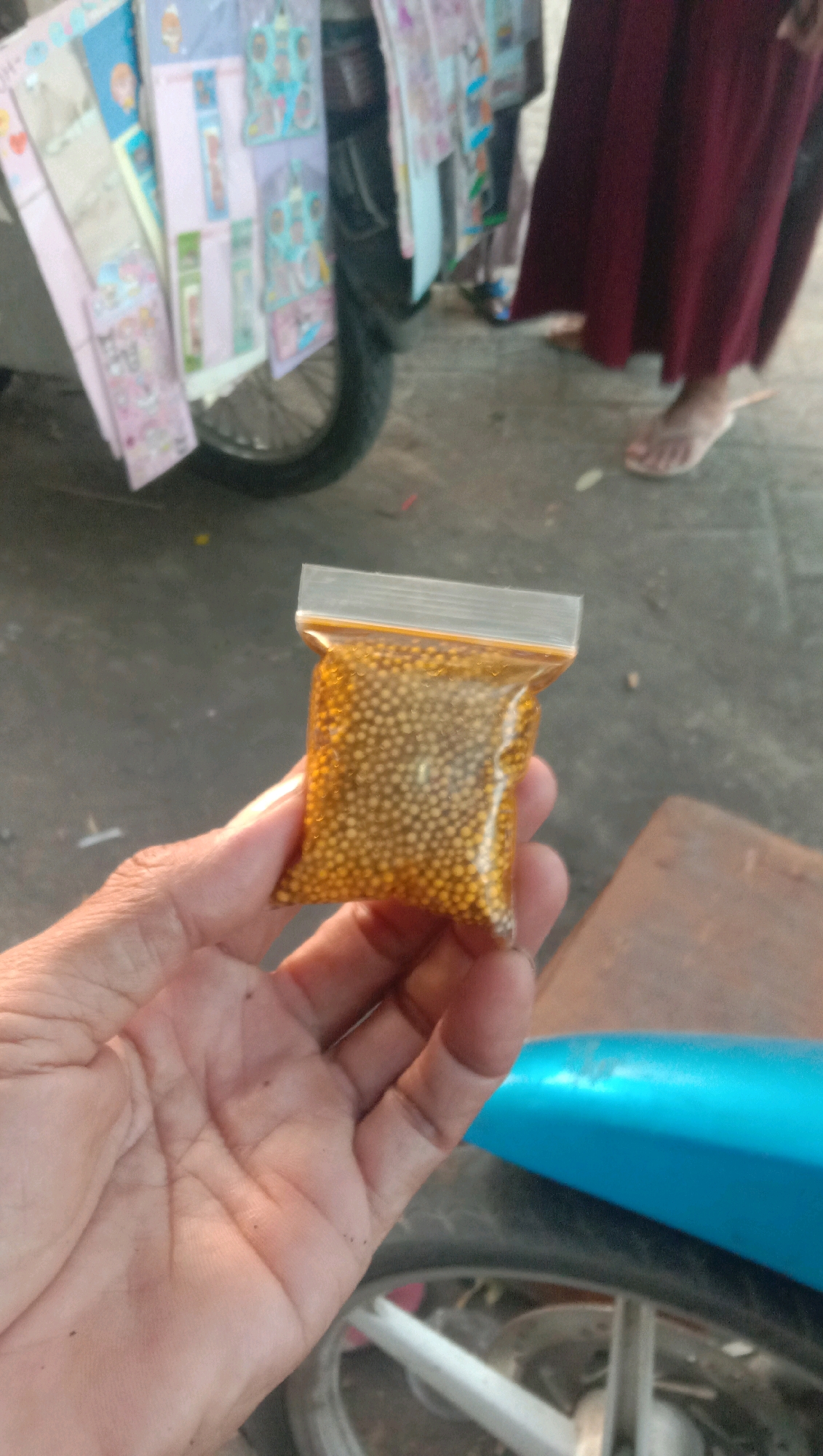 An adult's left hand is holding a small, clear plastic bag filled with tiny, golden beads. The bag is sealed at the top. The background is out of focus but appears to be an outdoor setting, possibly a street or market. A wheel of a vehicle, possibly a motorcycle, is visible in the lower part of the image. In the background, there is a glimpse of a person wearing a dark red garment and flip-flops, and another person's feet in blue sandals. There are also colorful posters or stickers attached to a surface in the background. The scene is set in Ngempit, Indonesia.