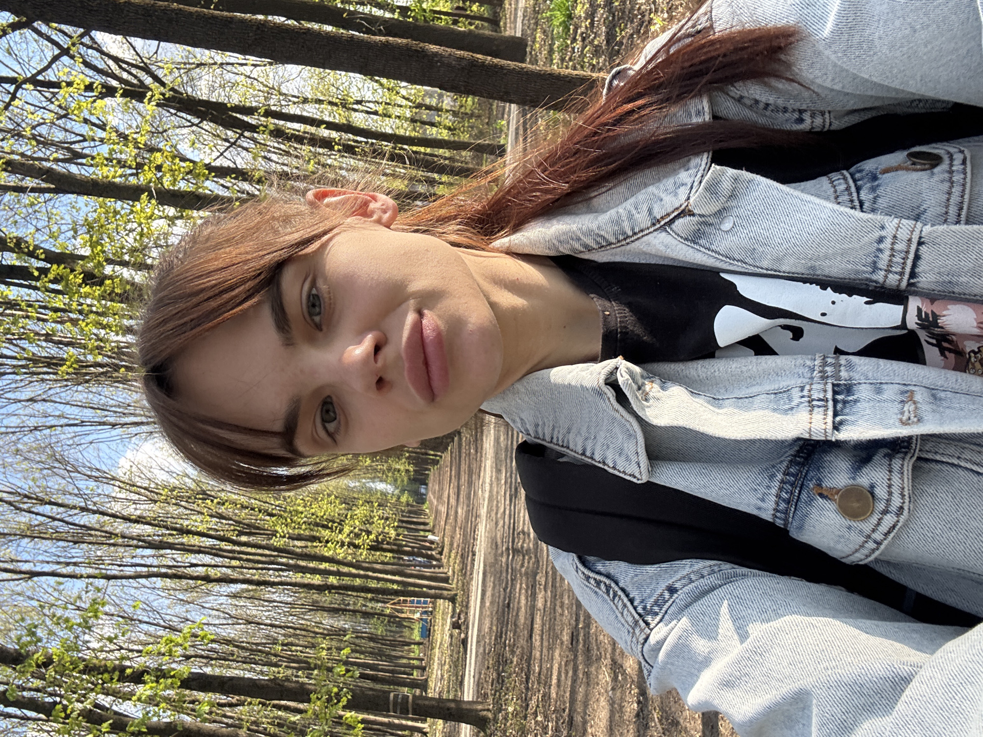 This media file presents a close-up, selfie-style portrait of a young person, likely female, captured outdoors in what appears to be a park or tree-lined avenue in Rostov-on-Don, Russia. The image is oriented with the subject's head tilted 90 degrees to the left. The subject has light brown hair, light eyes, and a neutral expression, looking directly at the camera. She is wearing a light-wash denim jacket over a black t-shirt that features a partial white graphic design on the front; the specific details or text of which are not fully discernible. A black strap, likely from a backpack, is visible over her right shoulder.
The background features a dense alignment of tall, slender trees flanking a wide dirt or gravel path, suggesting an early spring setting as new, small green leaves are visible on the branches. The sky is clear and blue, indicating a bright, sunny day. Strong sunlight illuminates the scene, casting shadows. The overall activity appears to be a casual moment during a stroll in a natural setting.