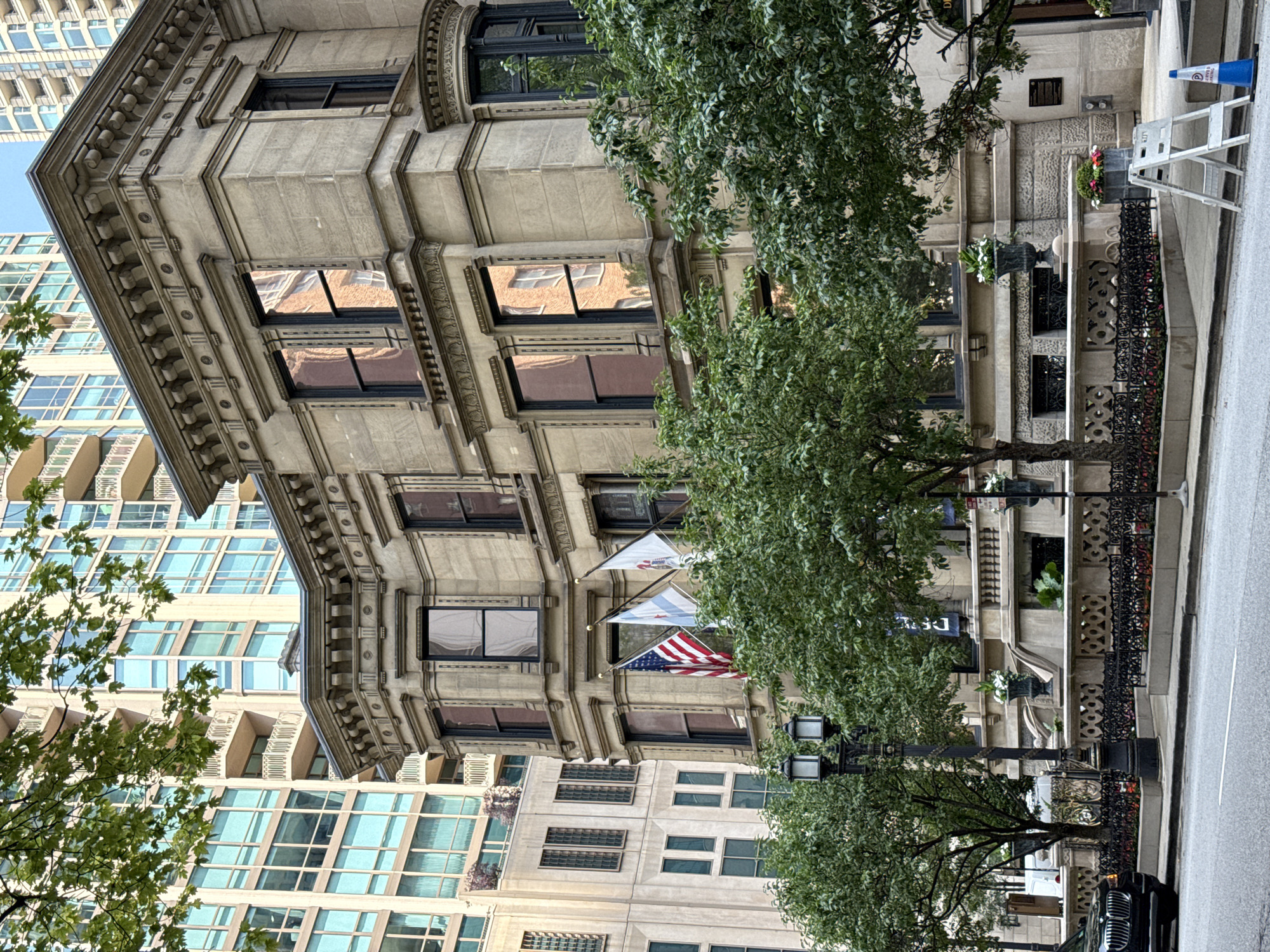 This media file captures a street view in Chicago, USA, featuring a mix of historic and modern architecture. The foreground is dominated by a vintage stone building with ornate detailing around its windows and eaves. Its facade is adorned with decorative molding and columns. Large trees with lush green foliage partially obscure the view of this building, casting shadows and adding a natural element to the urban scene.
Behind the historic structure, modern high-rise buildings with extensive glass facades are visible. These contemporary skyscrapers create a stark contrast with the older architecture. Three flags are displayed on poles attached to the stone building: an American flag and two other flags, one of which appears to be a state or city flag, possibly Illinois.
The street is paved, and a sidewalk is visible along the edge of the property. Several objects are present on the sidewalk, including a black metal fence with decorative scrollwork, planters with flowers, and a portable white ladder. A blue traffic cone and a white A-frame ladder suggest some form of outdoor work or event preparation. A black car is parked along the side of the road in the lower right corner.
The lighting suggests it is daytime, likely late afternoon, with the sun casting some shadows but not being intensely bright, indicating potentially overcast or partly cloudy weather. The scene conveys a sense of a bustling urban environment where historical preservation meets modern development. No people are prominently visible. There is no discernible text in the visible portion of the image.