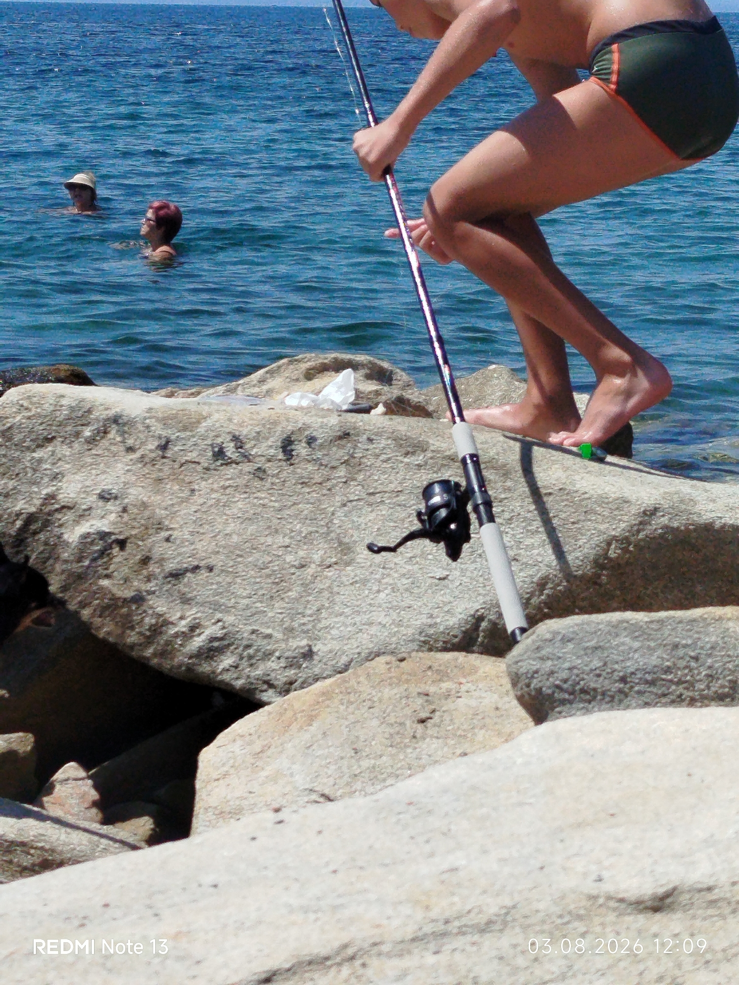 In Sithonia Municipal Unit, Greece, on a rocky shoreline, a person is fishing with a rod and reel, standing on large, light-colored boulders near the sea. The water is blue and appears somewhat choppy. In the background, two individuals are swimming or wading in the sea. One swimmer is wearing a hat, and the other has red hair. The foreground is dominated by the rocks and the fishing equipment. The image also contains text overlays indicating "REDMI Note 13" and "03.08.2026 12:09".