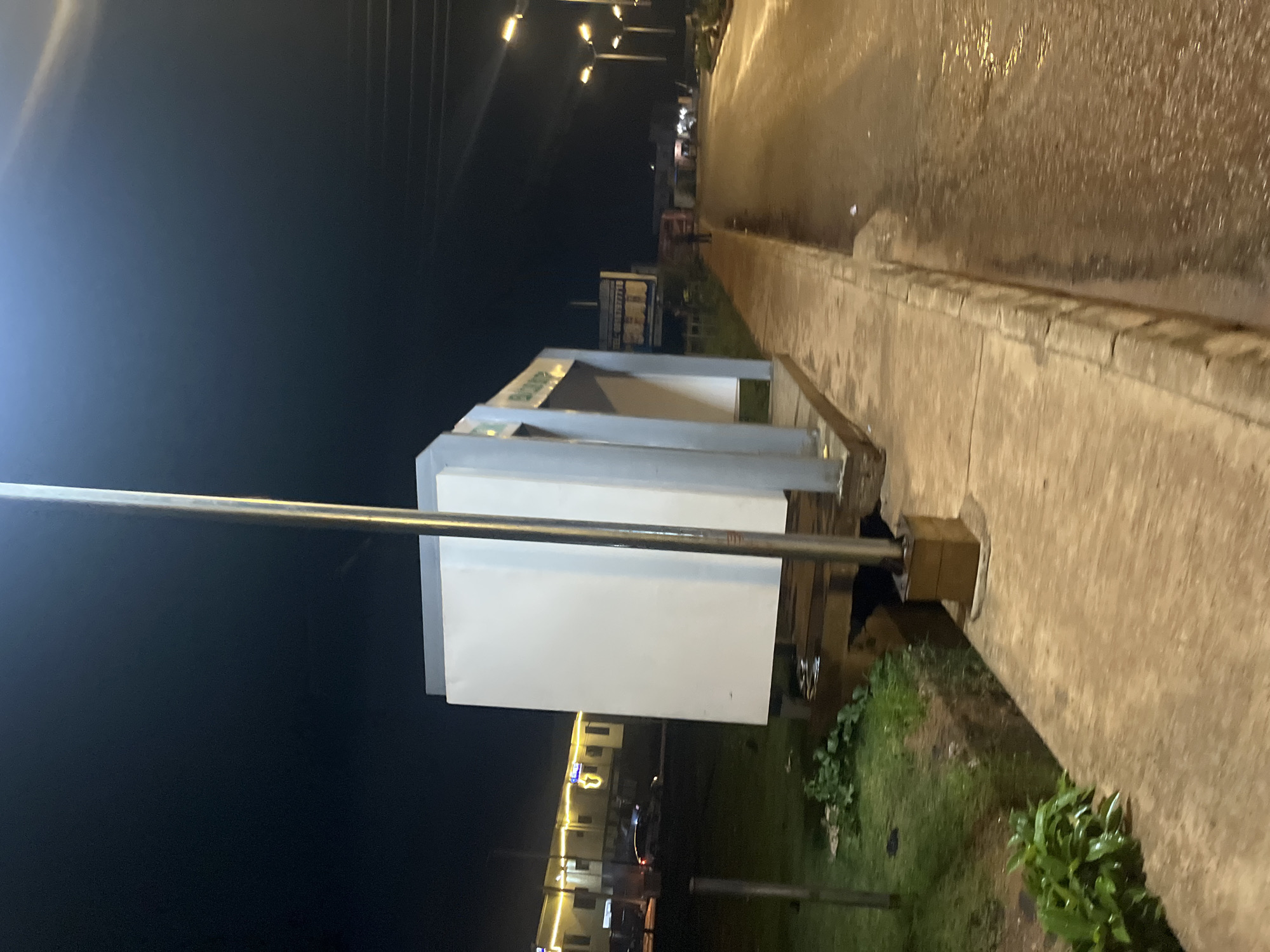 The image depicts a nighttime scene in Bukuru, Nigeria, focusing on a roadside structure that appears to be a bus stop or shelter. The structure is modern in design, with a predominantly white front panel and grey framing, standing on a concrete base. A tall, silver light pole is positioned prominently in front of the shelter, its light illuminating the immediate surroundings.
To the left of the shelter, there is a grassy area with some vegetation. Beyond the grass, buildings with yellow lighting are visible, suggesting commercial or residential properties. A dark-colored car is parked in front of these buildings.
The foreground consists of a concrete walkway or sidewalk that runs alongside the road. The road itself appears to be wet, reflecting the light from street lamps that line the right side of the image. The street lamps are modern, with multiple bulbs, casting a warm, yellow glow. Further down the road, other buildings and street lights are visible in the distance, indicating a developed urban area. A lone figure is standing on the sidewalk in the distance, facing away from the camera. The overall atmosphere is one of a quiet, illuminated night in a Nigerian city.