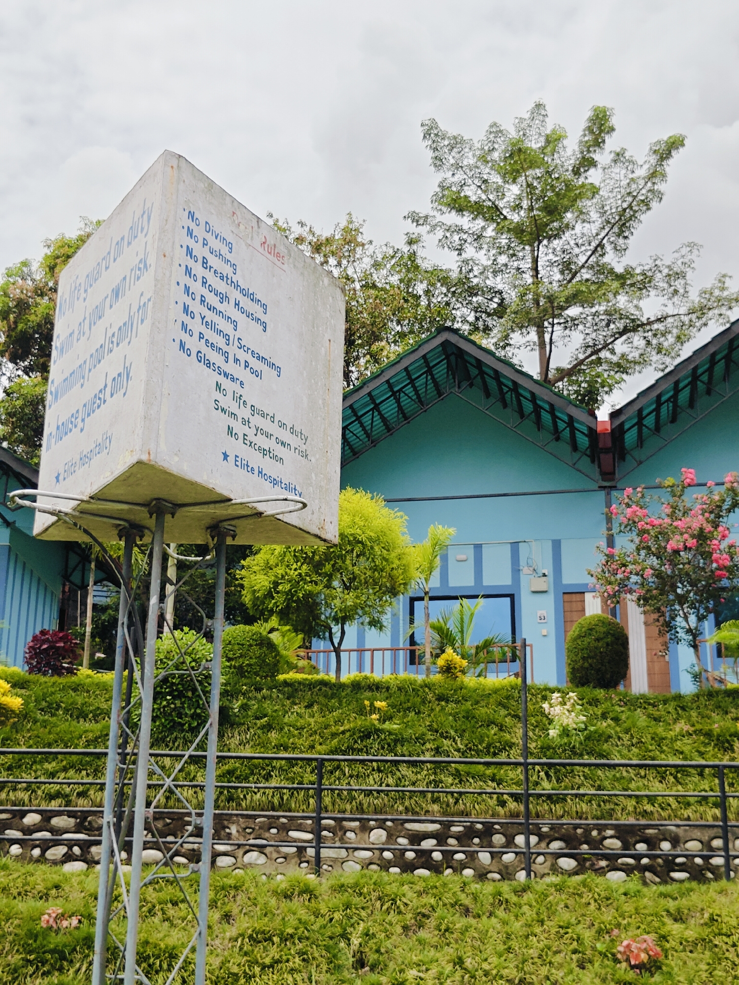 The image shows a close-up of a concrete sign with "Pool Rules" written in red at the top right corner of the sign. Below this, a list of rules is written in blue text. The rules include "No Diving," "No Pushing," "No Breathholding," "No Rough Housing," "No Running," "No Yelling/Screaming," "No Peeing In Pool," and "No Glassware." Further down, it says "No life guard on duty," "Swim at your own risk," and "No Exception." At the very bottom, it says "Elite Hospitality" with a blue star next to it.
The sign is mounted on a metal pole structure and is positioned in front of a light blue building with a green roof. The building appears to be a resort or hotel, given the landscaping and signage. In the background, there are trees and a cloudy sky. The foreground consists of lush green grass, bushes, and a decorative stone wall with a black metal fence. There are also some flowering plants and trees visible. The overall scene depicts an outdoor area, likely a swimming pool area, at a hospitality establishment in the city of Ichchhakamana, Nepal.