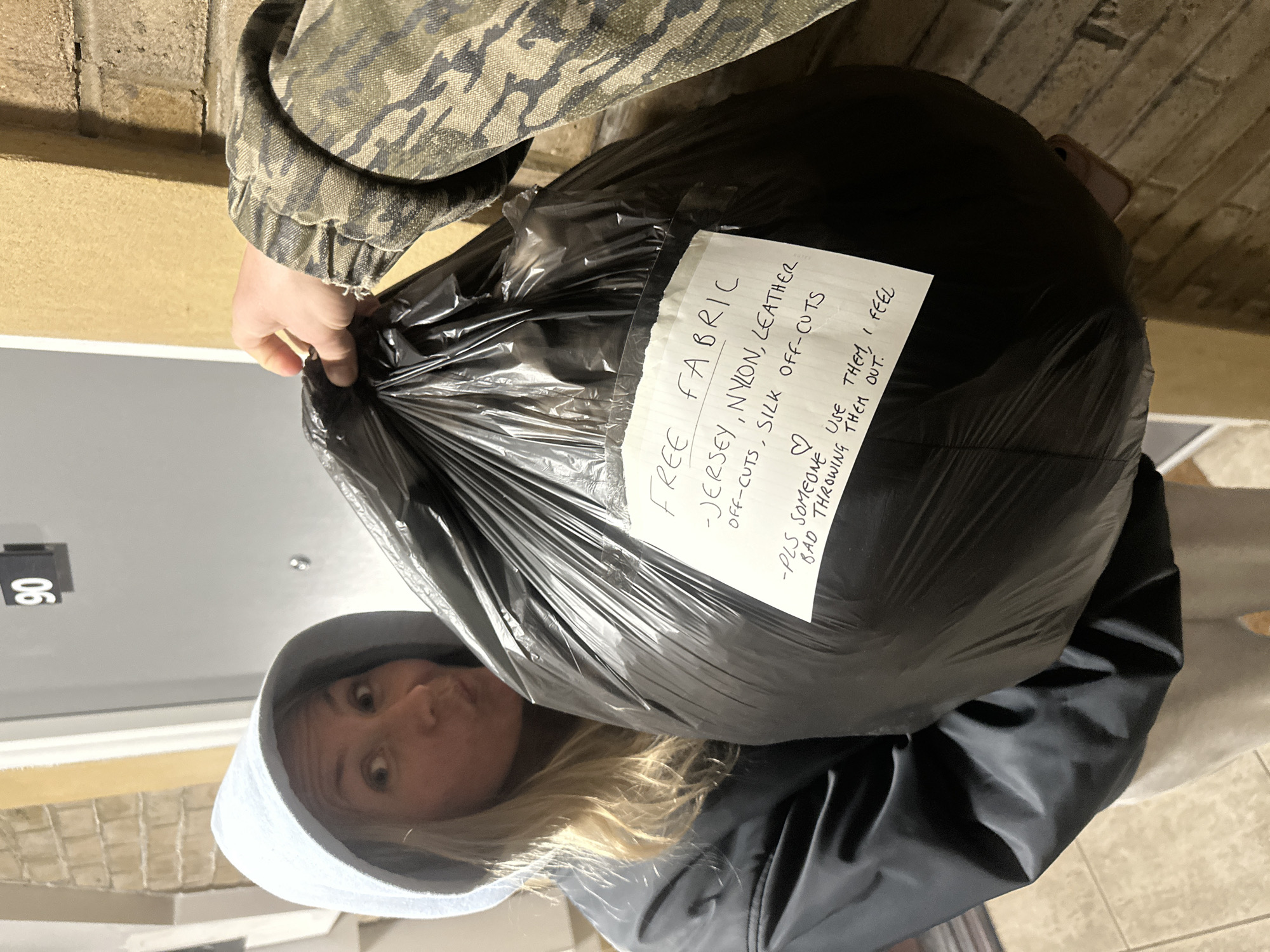 A woman in a grey hooded sweatshirt and black jacket is holding up a large black plastic bag filled with fabric offcuts. The bag has a handwritten note attached to it that says "FREE FABRIC - JERSEY, NYLON, LEATHER OFF-CUTS, SILK OFF-CUTS -PLS SOMEONE USE THEM, I FEEL BAD THROWING THEM OUT." The woman is looking at the camera with a slight frown. A person wearing a camouflage jacket is holding the bag from above. The scene is set in front of a building entrance, which appears to be a residence or apartment building. The number "90" is visible on the building exterior. The location is not specified, however, the city is mentioned.